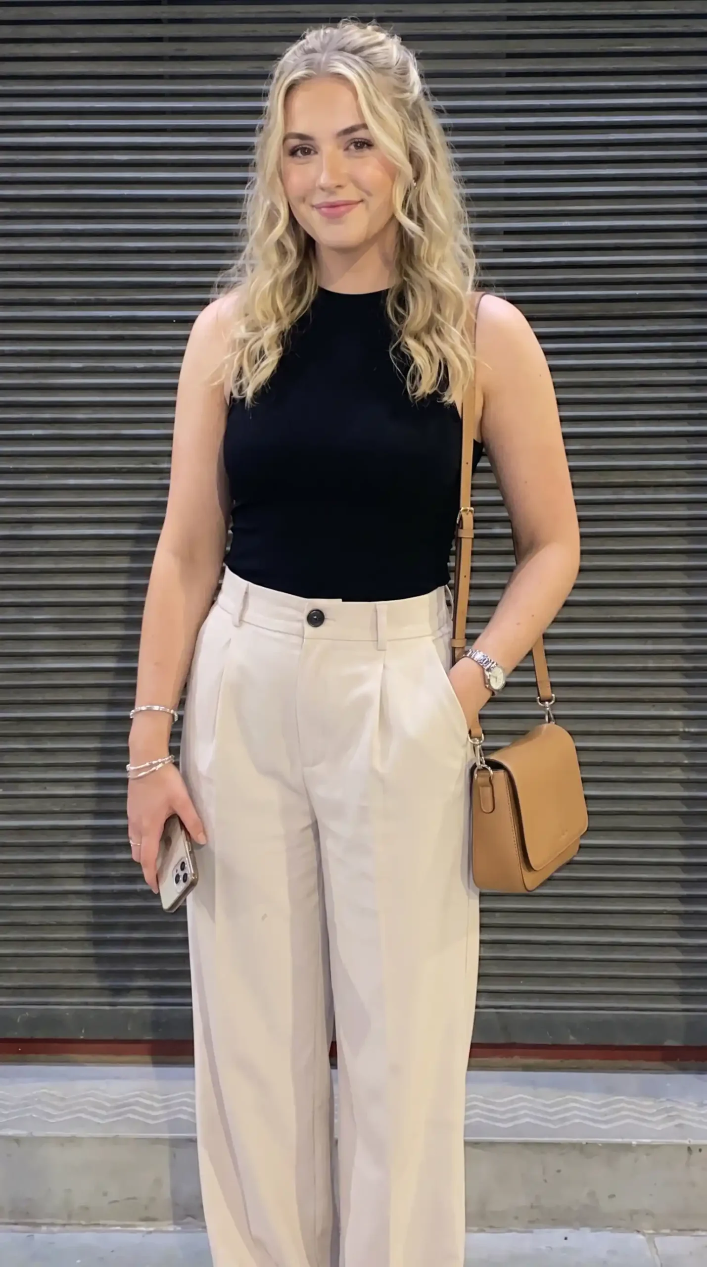 Woman wearing black racerback top with cream pleated trousers and tan crossbody bag