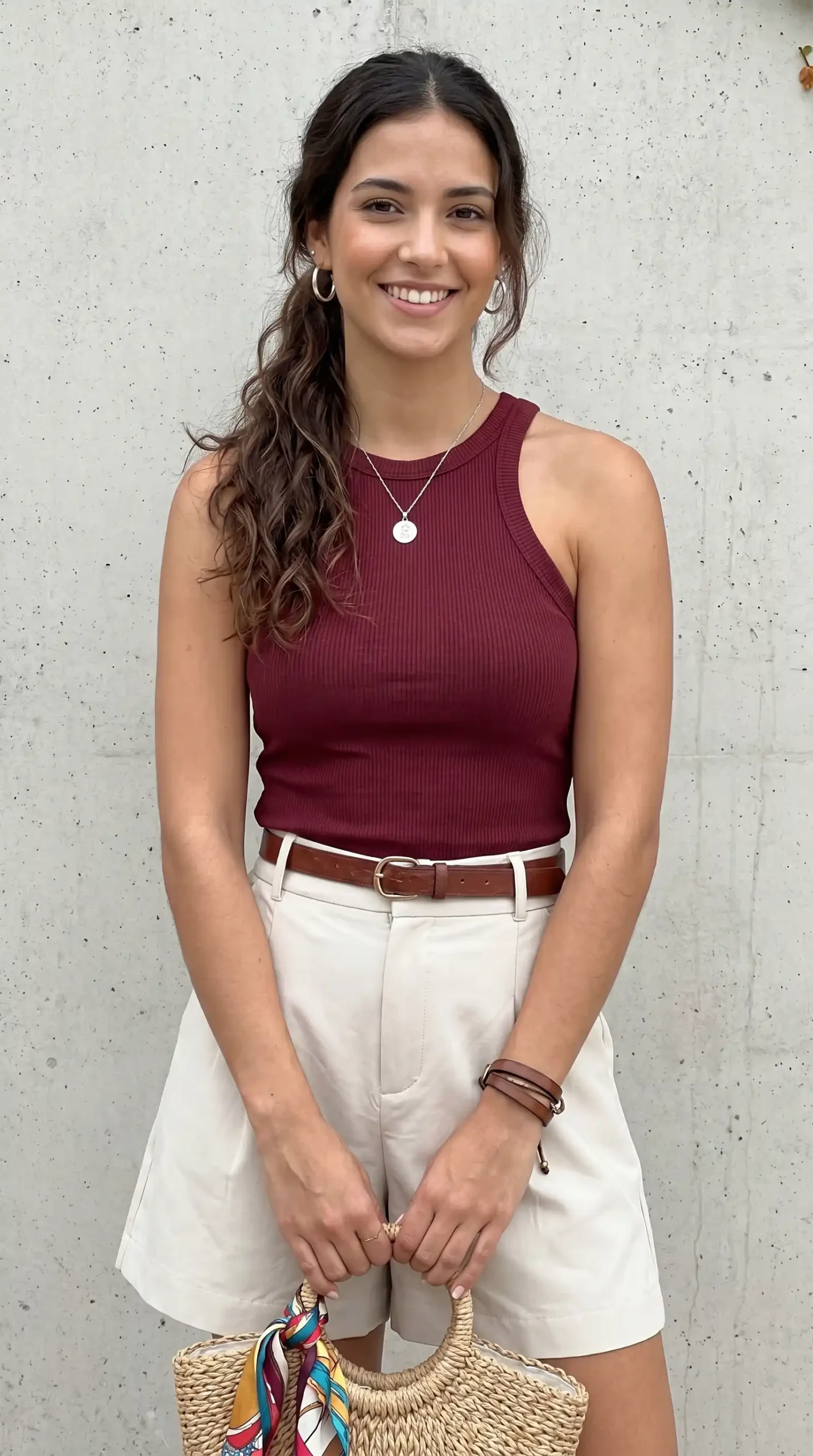 Woman wearing burgundy ribbed tank top with beige shorts and brown belt holding woven bag