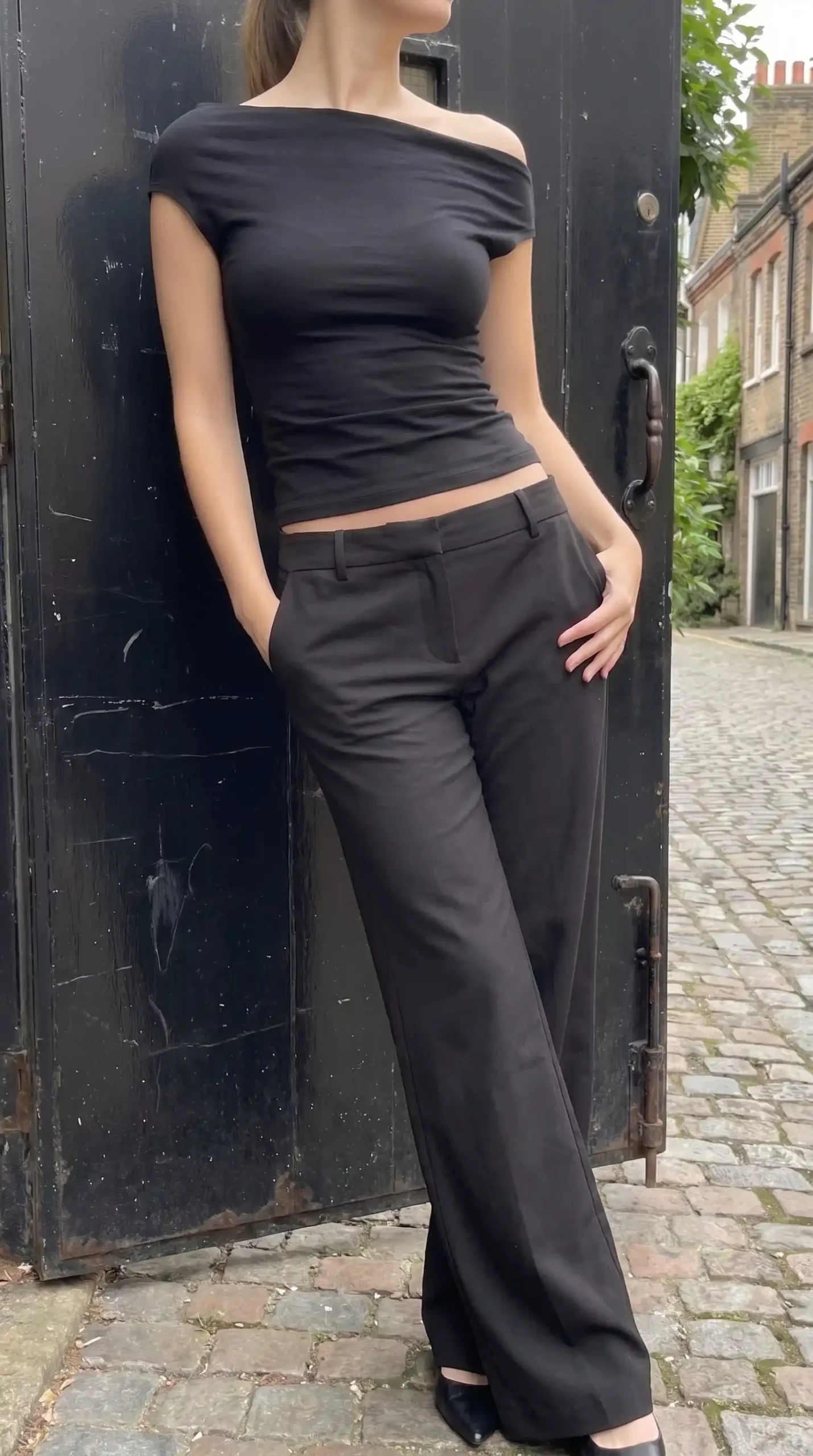 Woman in black off shoulder asymmetric draped crop top with black low rise wide leg trousers on cobblestone street