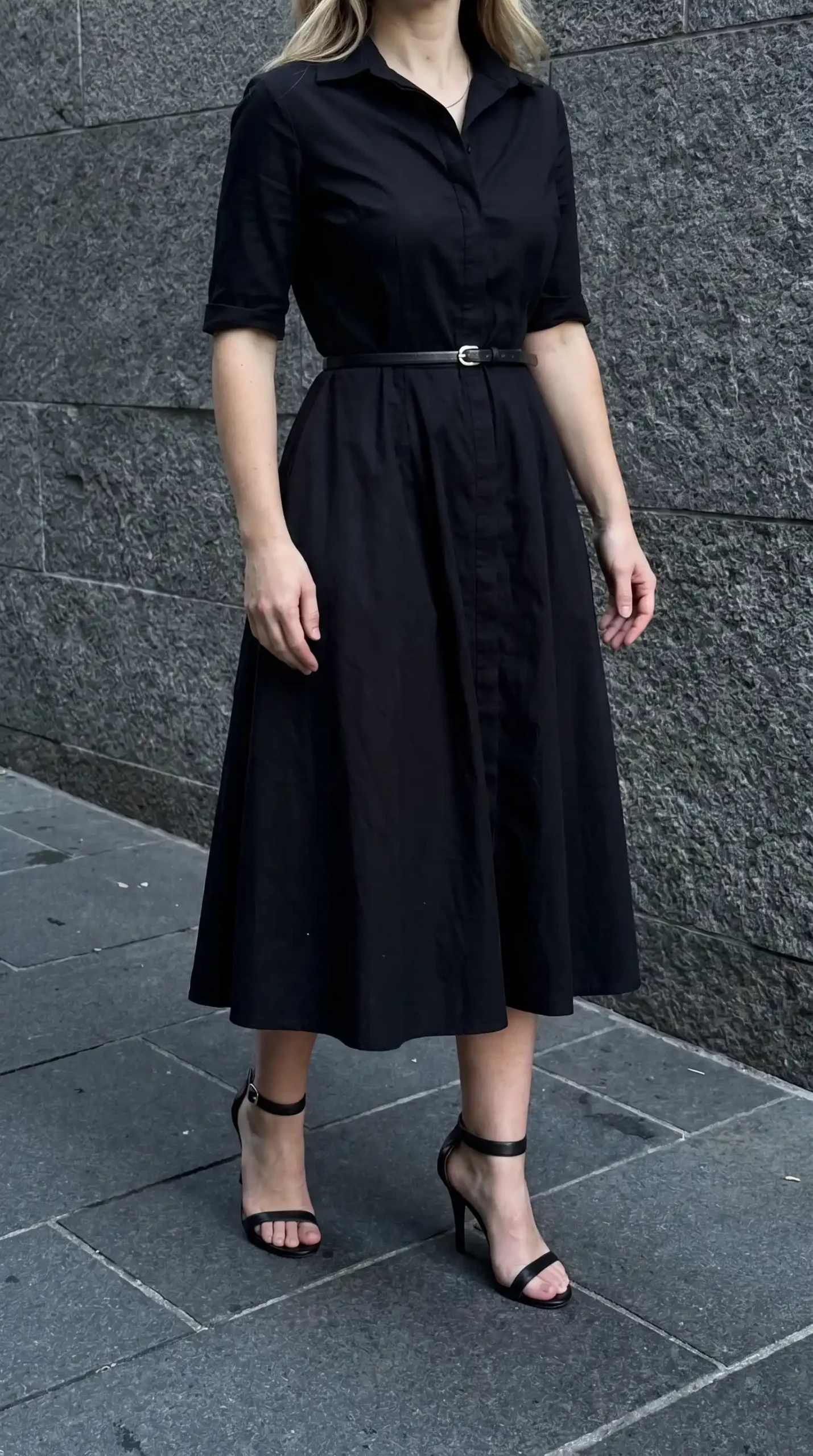 Woman in black short sleeve belted shirt midi dress with black strappy heeled sandals on cobblestone street