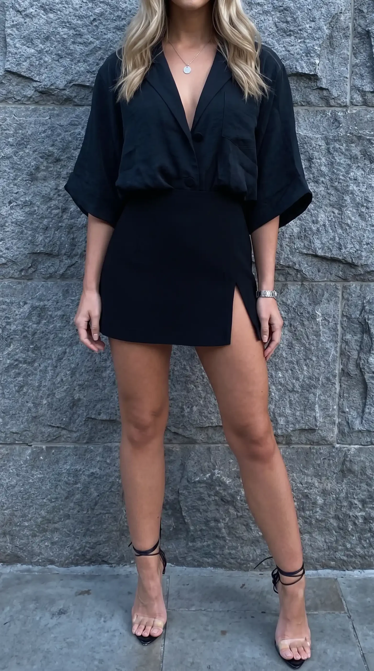 Woman in black deep v kimono sleeve blazer mini dress with black strappy heels against stone wall