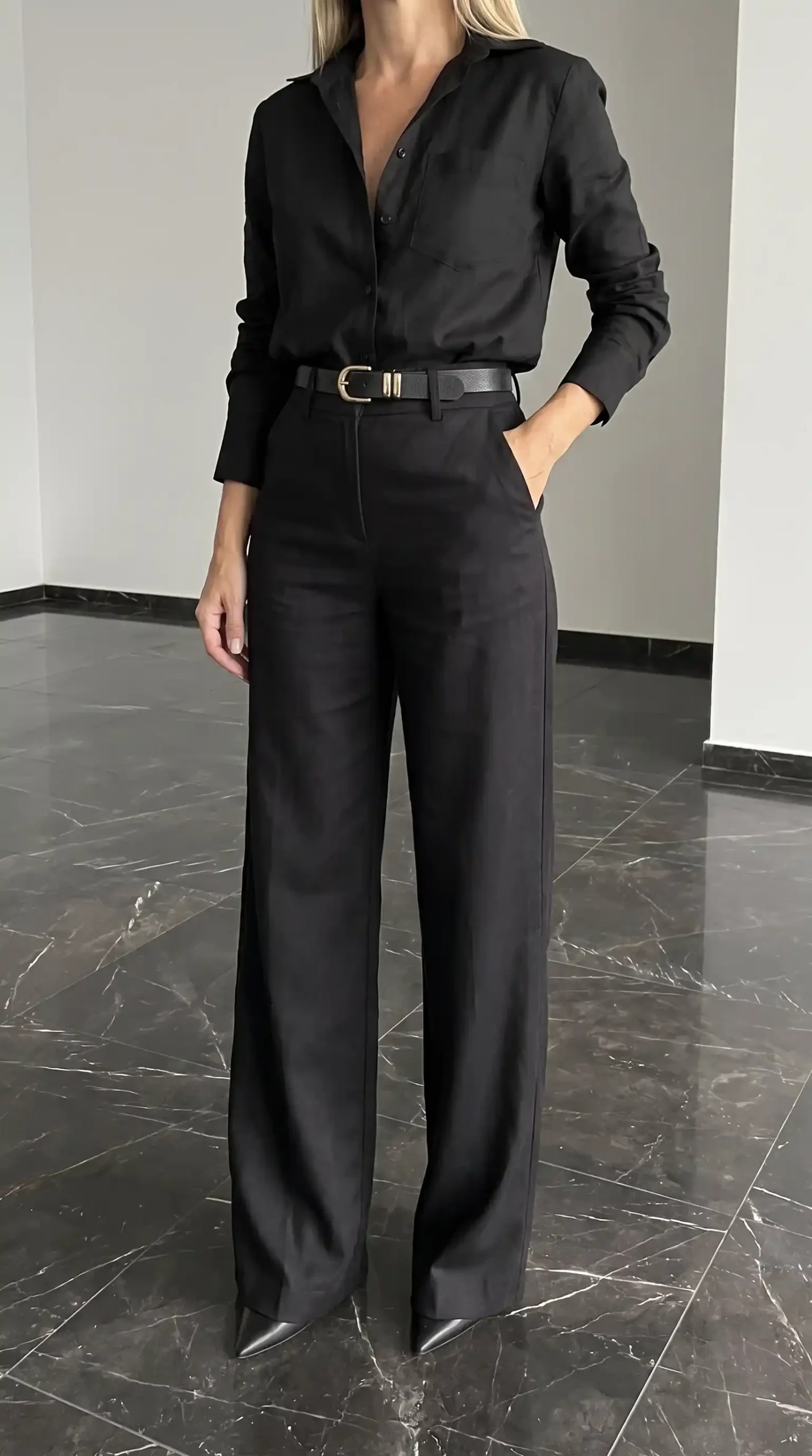 Woman in black linen button up shirt with black wide leg trousers and leather belt with gold buckle on marble floor