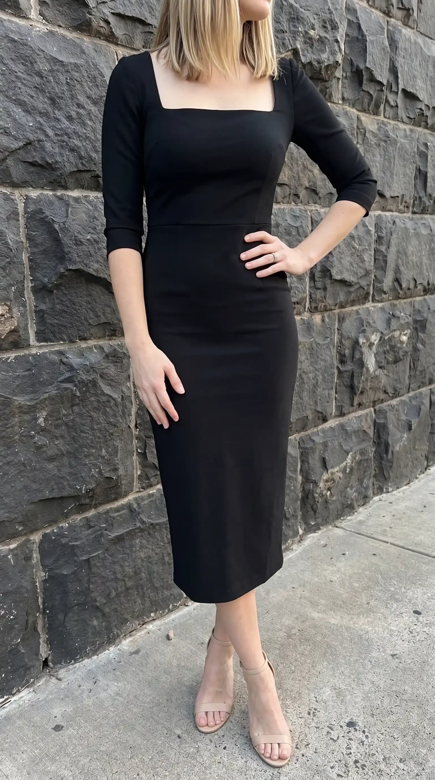 Woman in black square neck three quarter sleeve bodycon midi dress with nude strappy heels against stone wall
