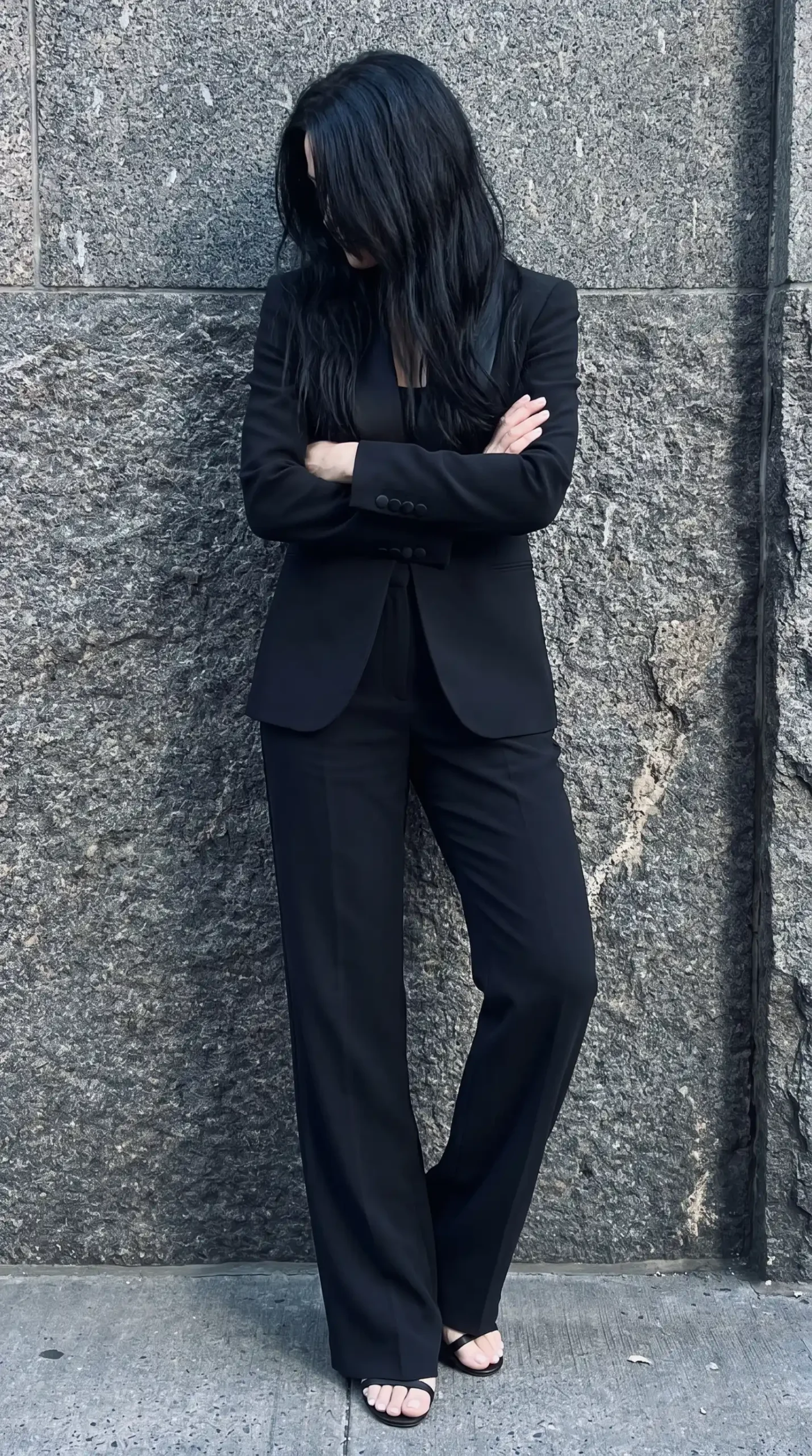 Woman in black tailored blazer with black camisole and wide leg tailored trousers against stone wall