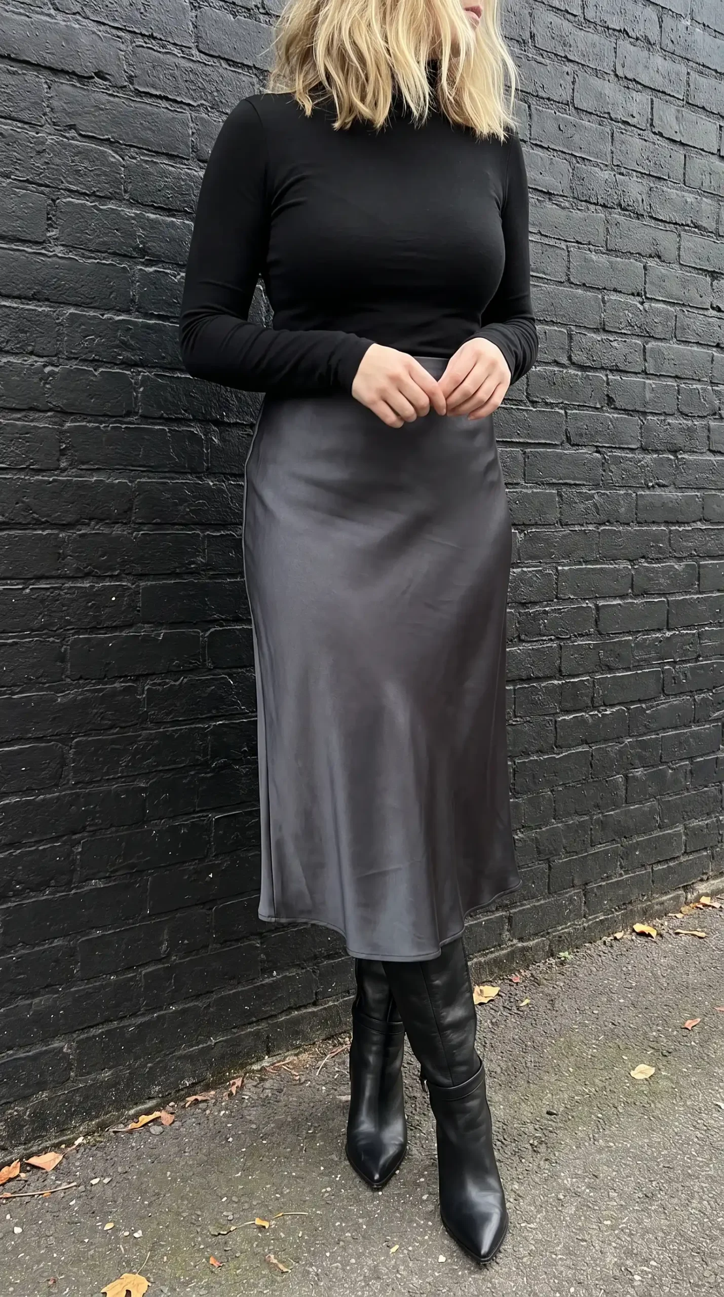 Woman in black turtleneck top with charcoal satin bias cut midi skirt and black knee high boots against brick wall