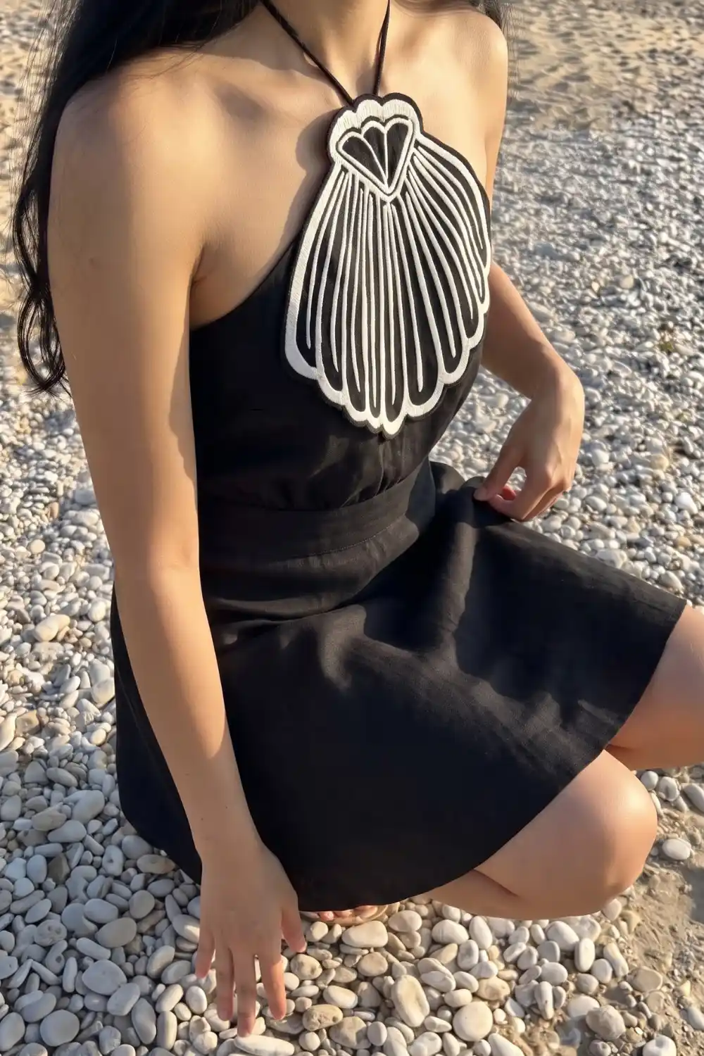 Woman in black halter mini dress with white shell embroidered bust on pebble beach