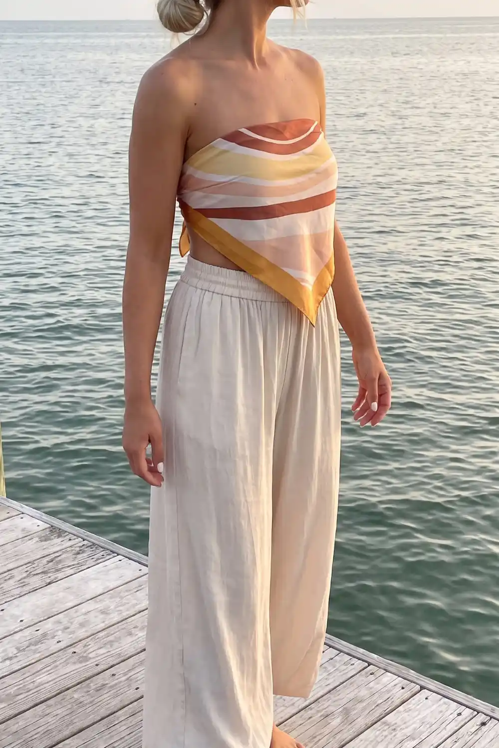 Woman in rust yellow stripe satin scarf strapless top with sand linen wide leg pants on wooden dock