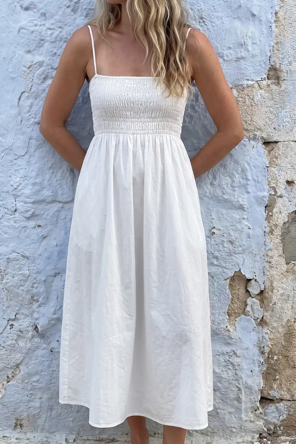 Woman in white shirred bodice spaghetti strap midi dress against blue whitewashed wall