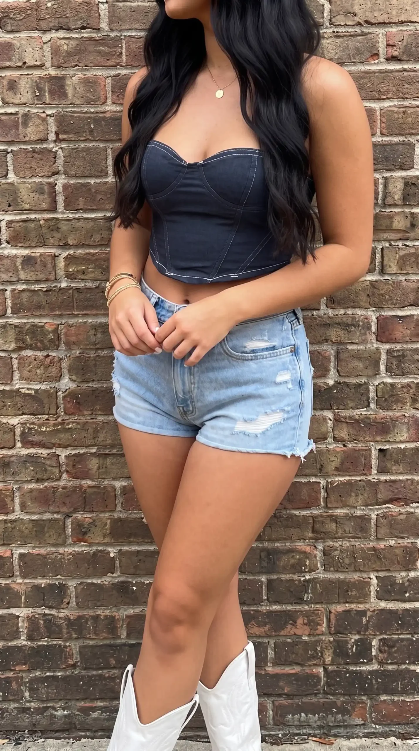 Woman in dark denim corset top with light wash ripped shorts and white cowboy boots against brick wall