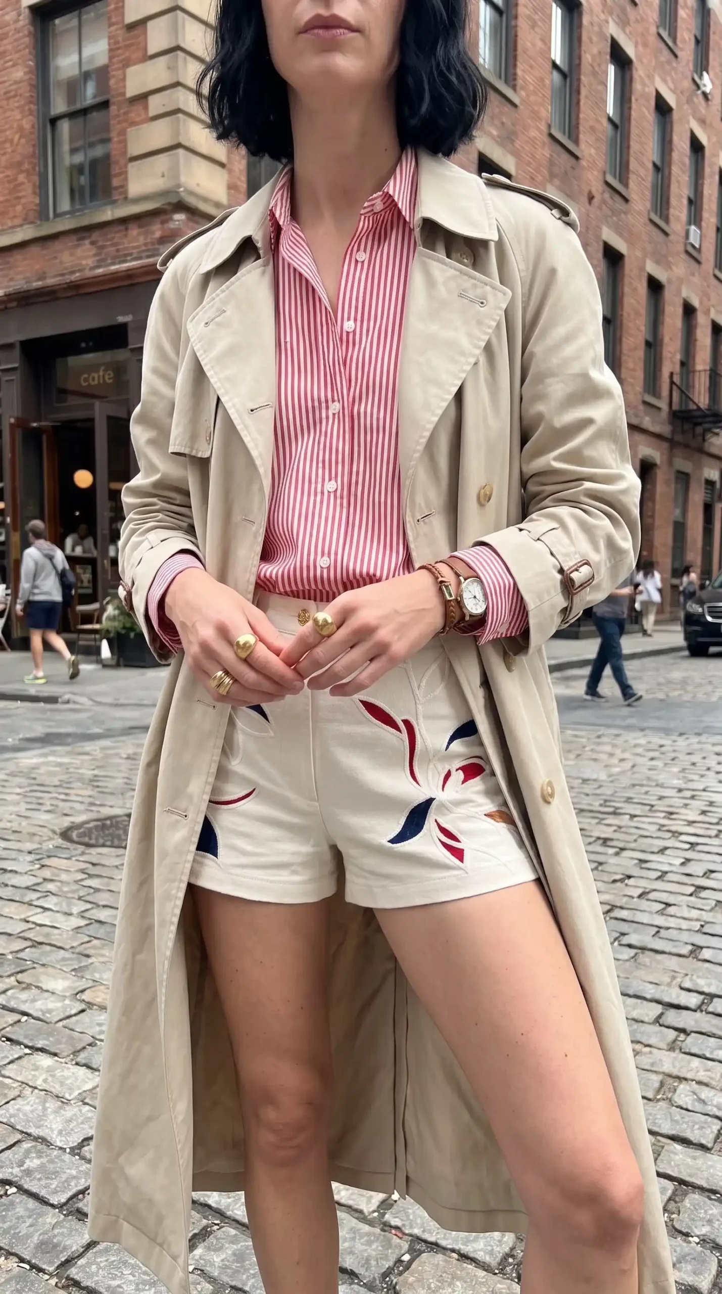 Woman wearing beige trench coat with pink striped shirt and cream floral embroidered shorts on cobblestone street
