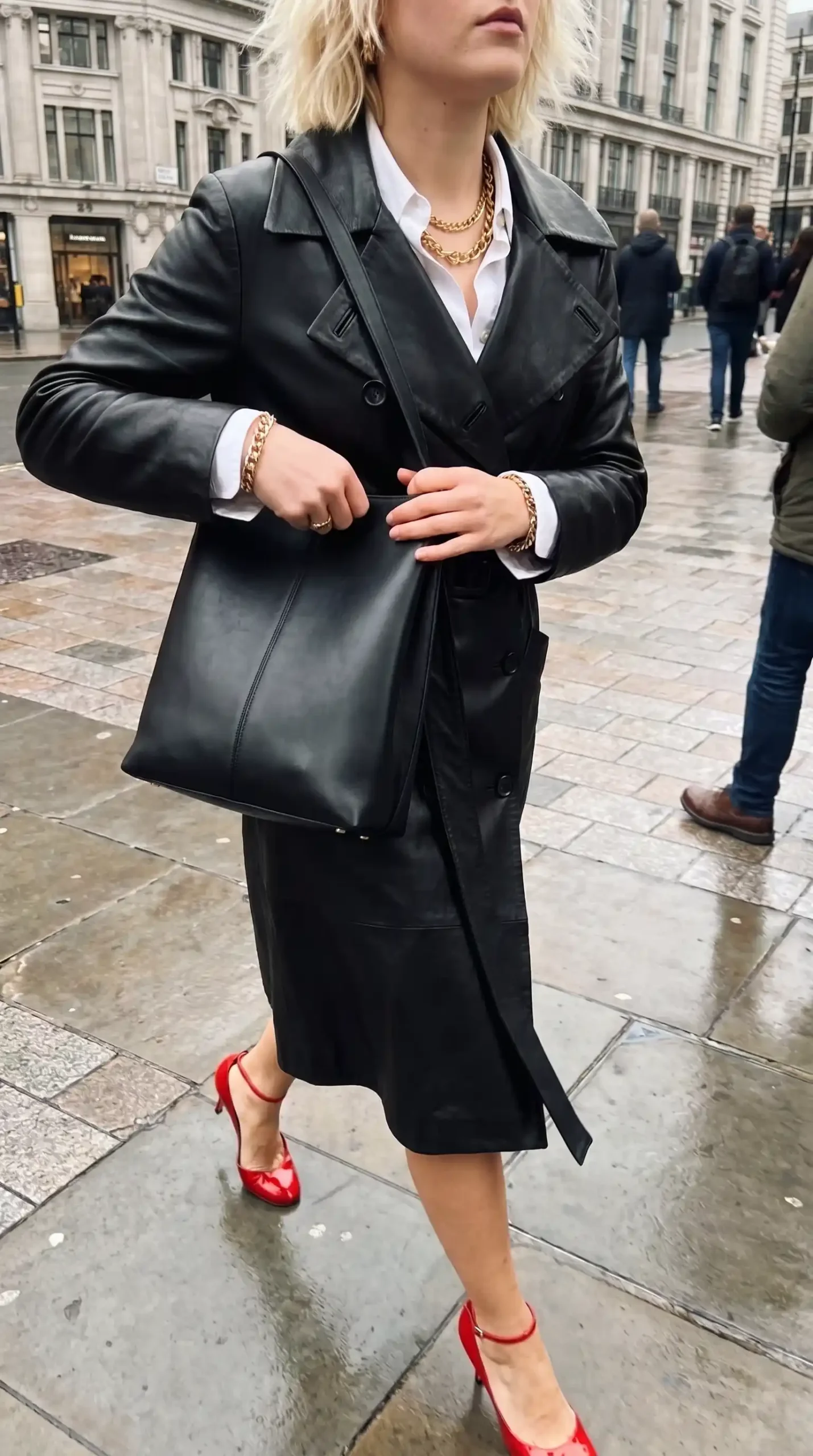 Woman wearing black leather double breasted trench coat with white shirt black leather midi skirt red mary jane heels and gold chain necklace on rainy London street
