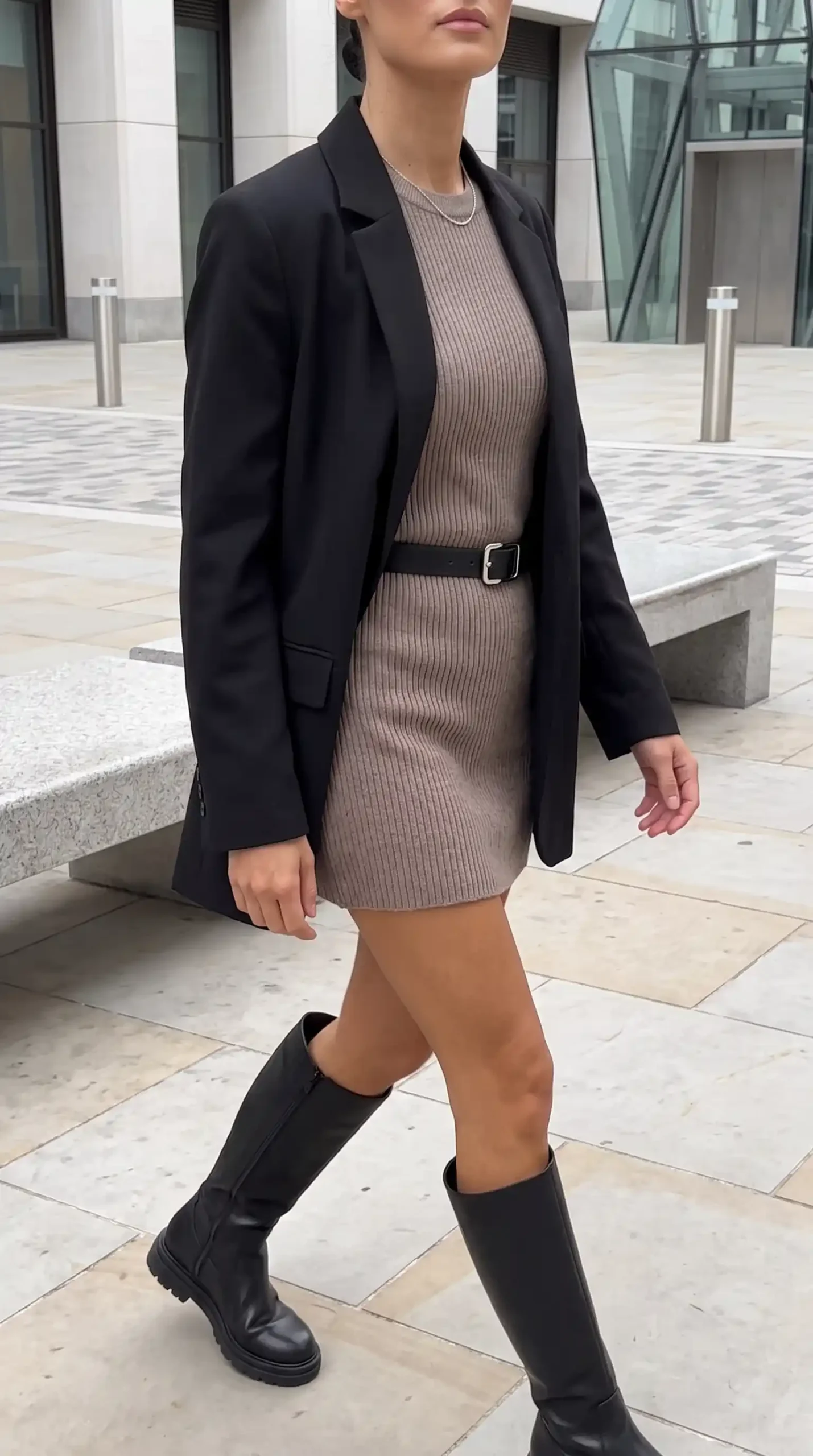 Woman wearing black oversized blazer over taupe ribbed knit mini dress with black belt and black knee high boots outdoors