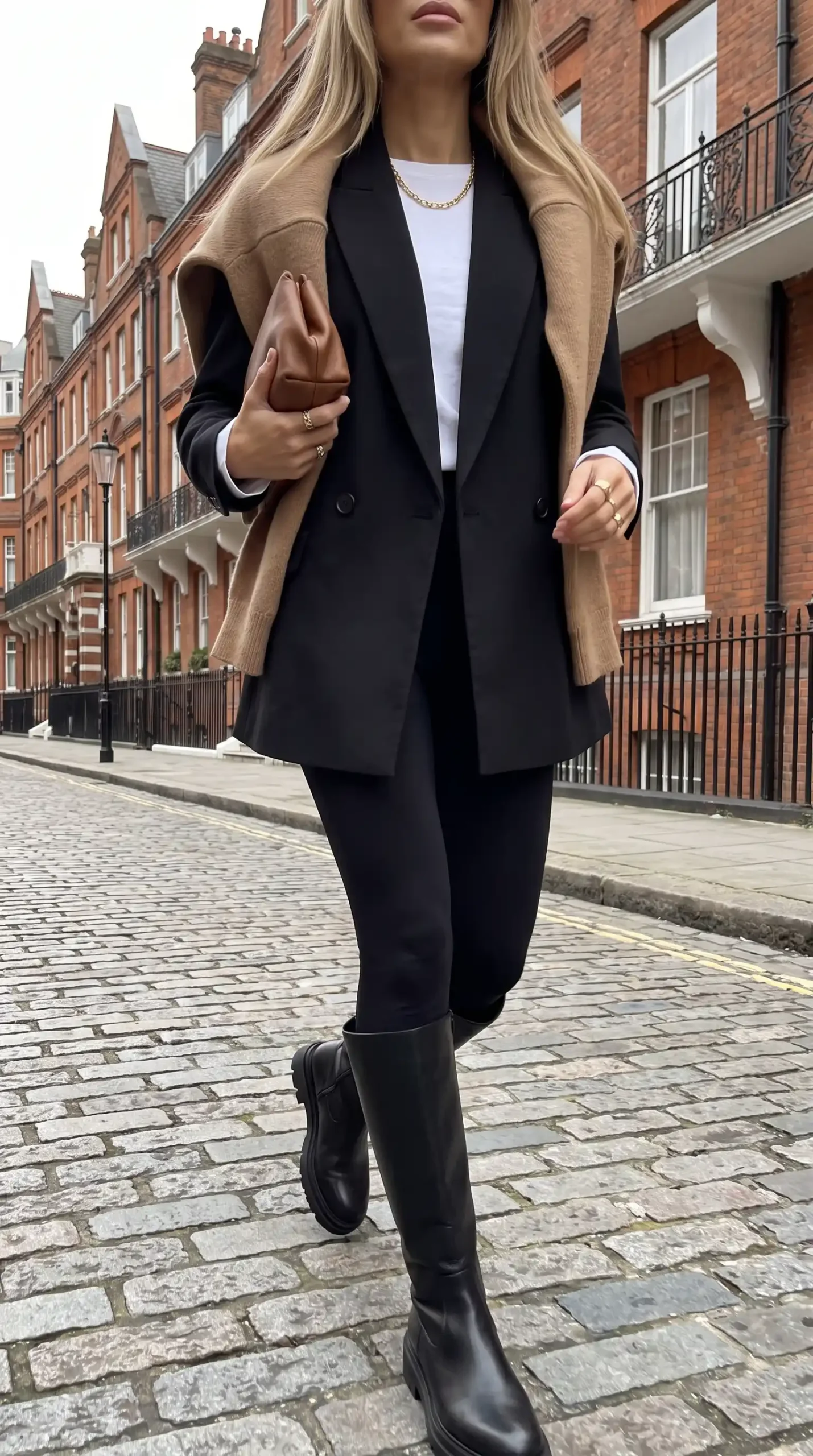 Woman wearing black oversized blazer with white tee black leggings camel knit draped over shoulders and black knee high boots on cobblestone street