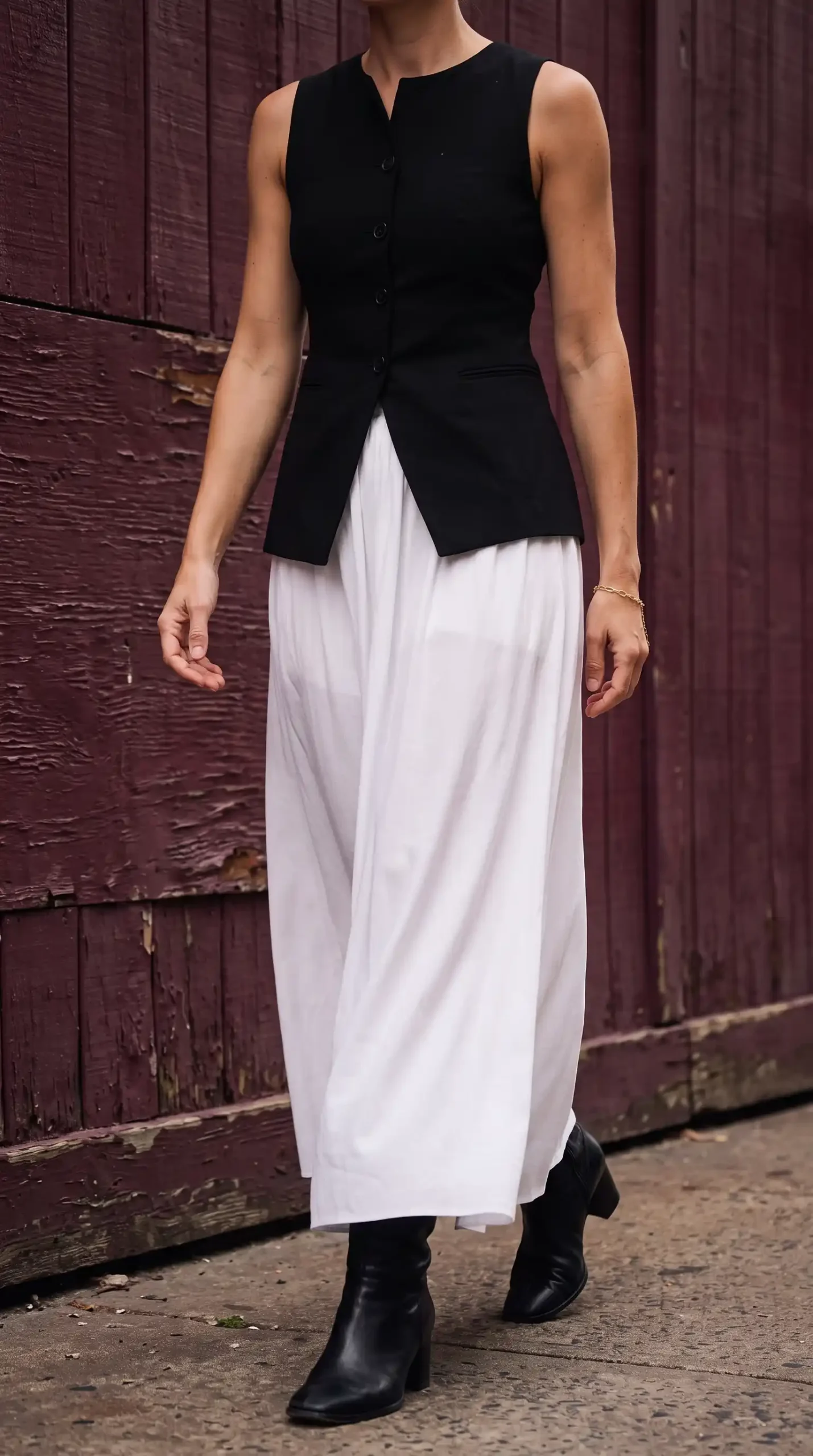 Woman in black button front vest with white flowy maxi skirt and black ankle boots against red wooden wall
