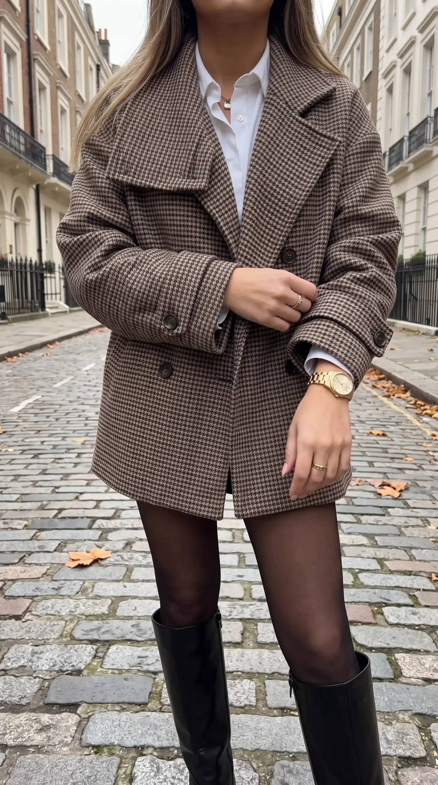 Woman wearing brown houndstooth oversized wool coat with white shirt black sheer tights and black knee high boots on cobblestone street