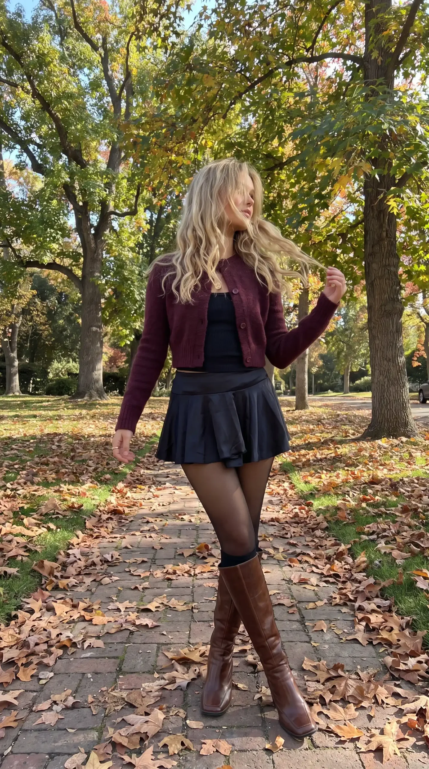 Woman wearing burgundy cropped cardigan with black crop top black faux leather pleated mini skirt sheer tights and brown knee high boots in autumn park