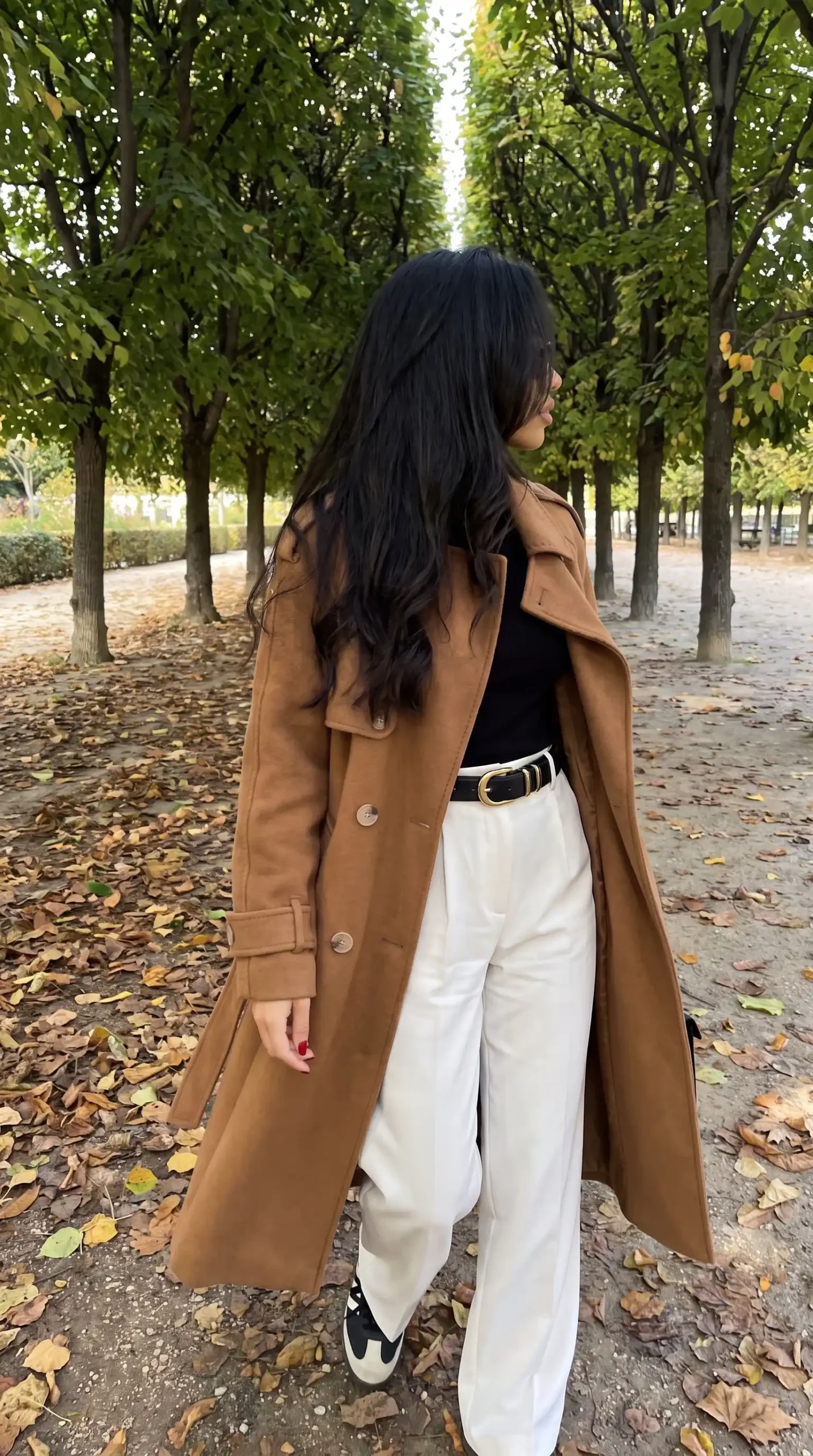Woman wearing camel wool trench coat with black top white wide leg trousers gold belt and black white sneakers in autumn park
