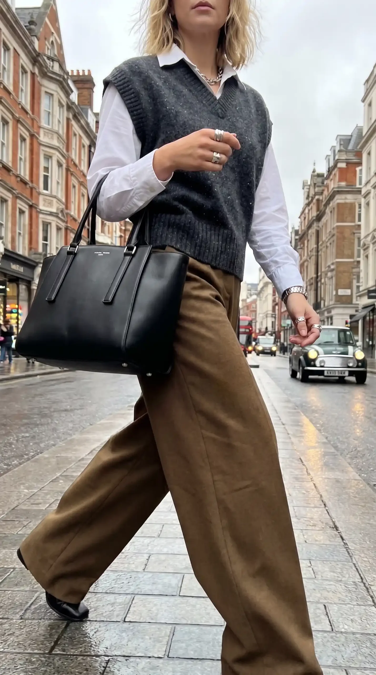 Woman wearing charcoal grey knit vest over white shirt with khaki wide leg trousers and black leather tote on wet London street