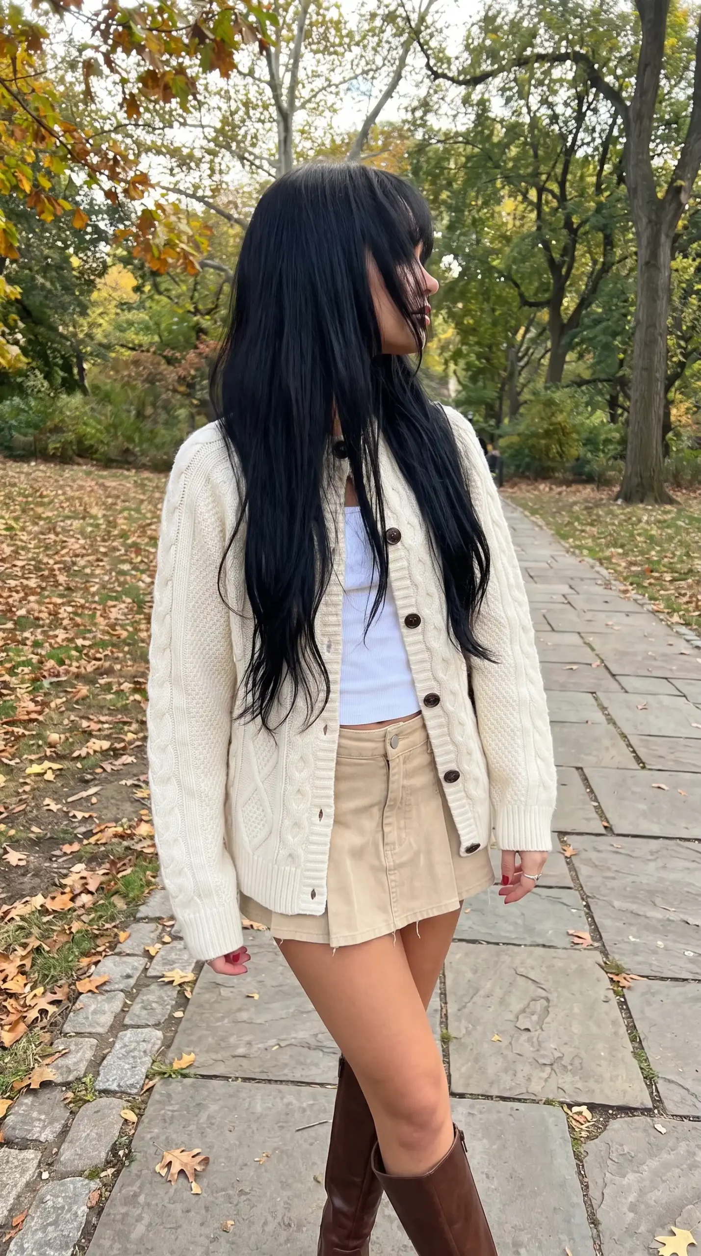 Woman wearing cream cable knit cardigan with white ribbed crop top khaki mini skirt and dark brown knee high boots in autumn park