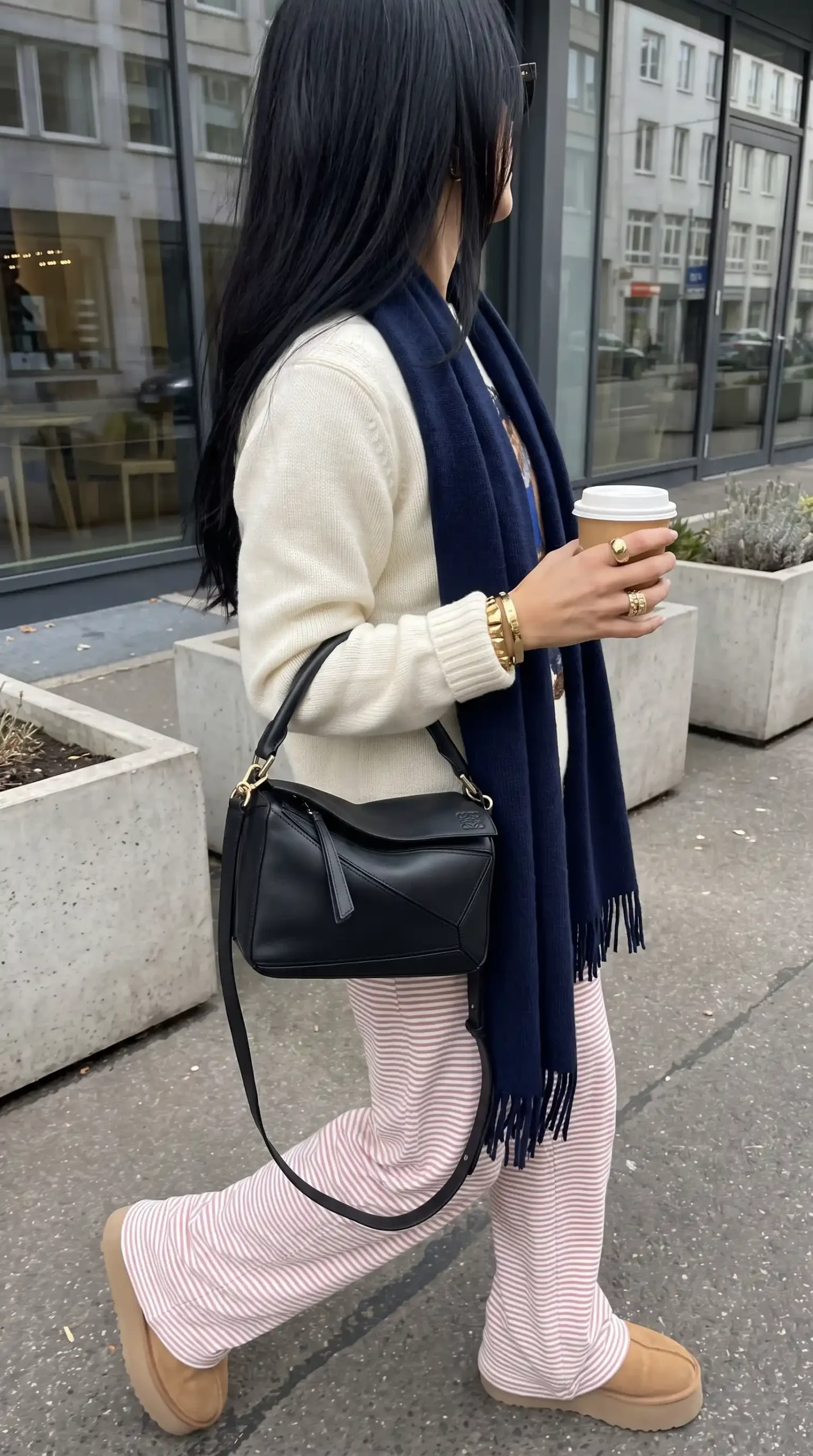 Woman wearing cream knit sweater with pink striped wide leg trousers navy fringe scarf black crossbody bag and tan Ugg boots outdoors