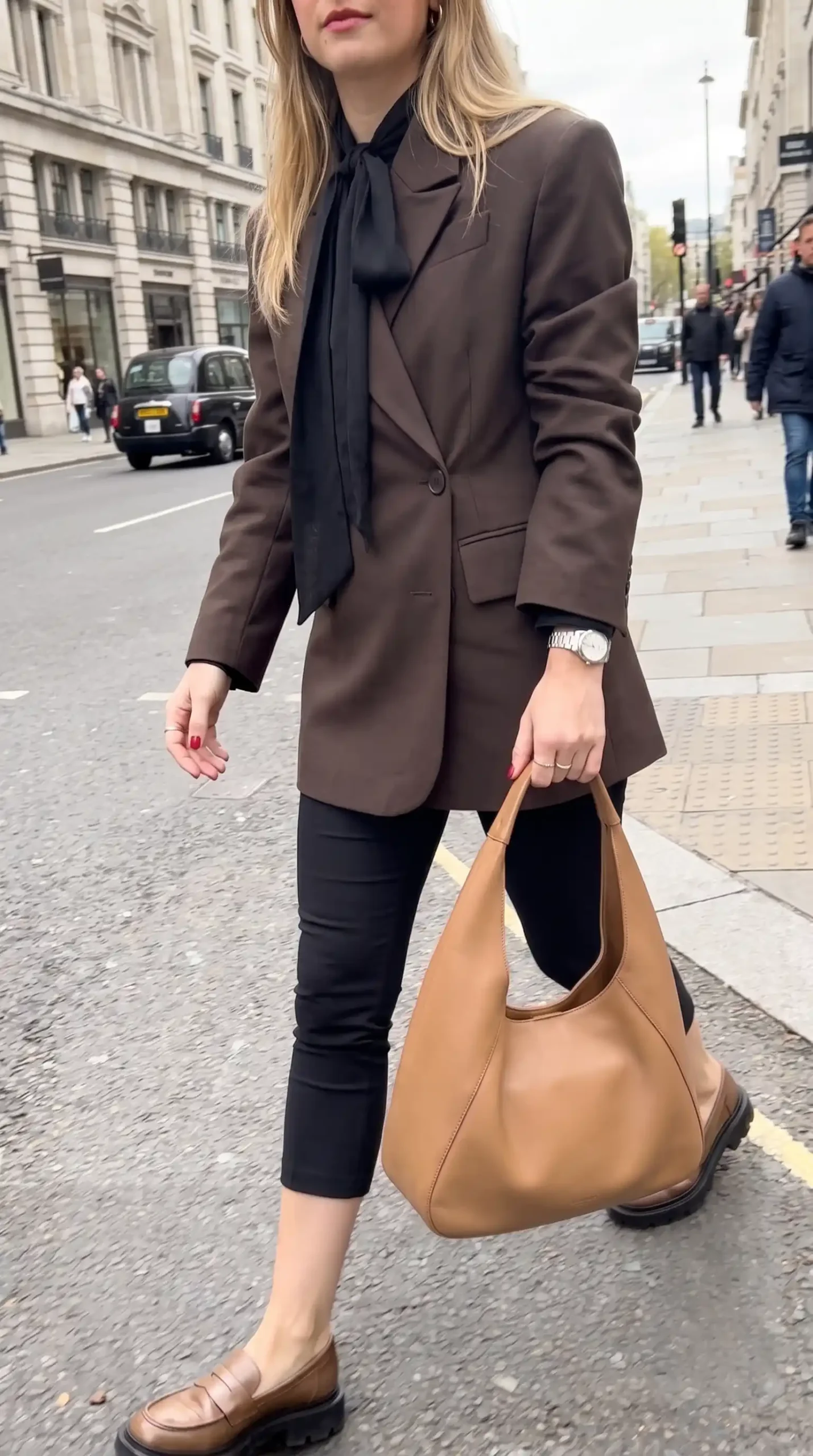 Woman wearing dark brown oversized blazer with black pussy bow blouse black skinny jeans tan hobo bag and brown chunky loafers on London street