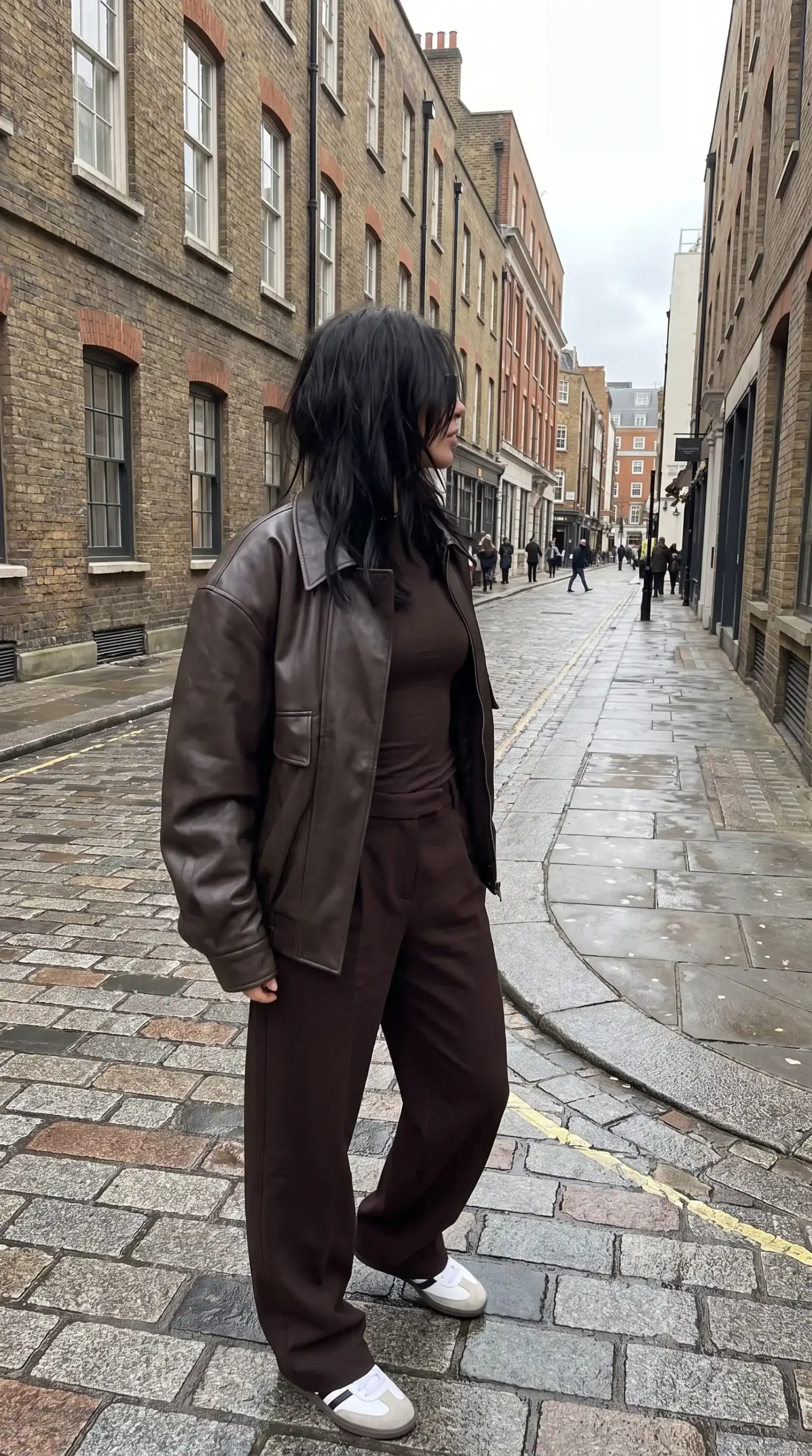 Woman wearing dark brown oversized leather jacket with brown turtleneck and wide leg trousers and white sneakers on wet cobblestone street