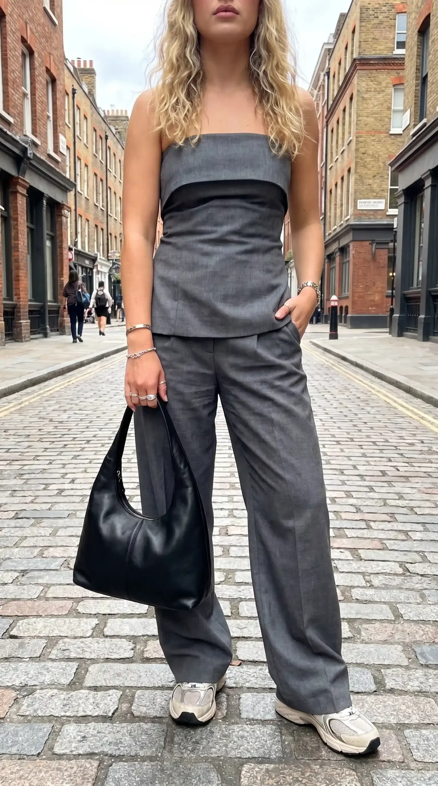 Woman wearing grey tailored strapless corset top with matching wide leg trousers black leather shoulder bag and white chunky sneakers on cobblestone