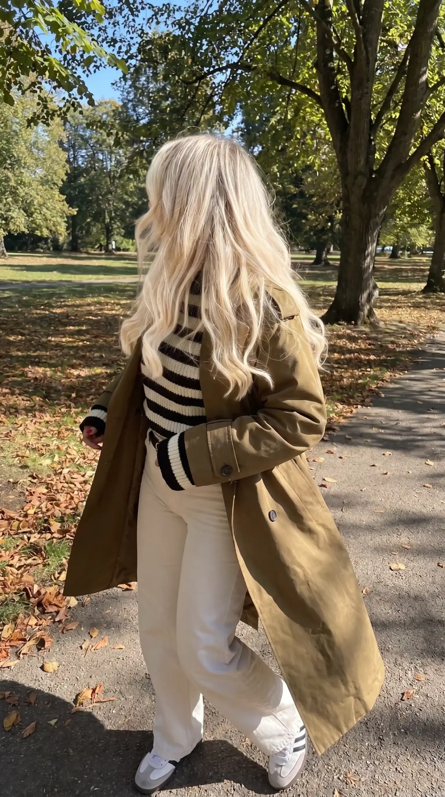 Woman wearing khaki olive trench coat with cream and black striped knit sweater cream wide leg trousers and white sneakers in autumn park