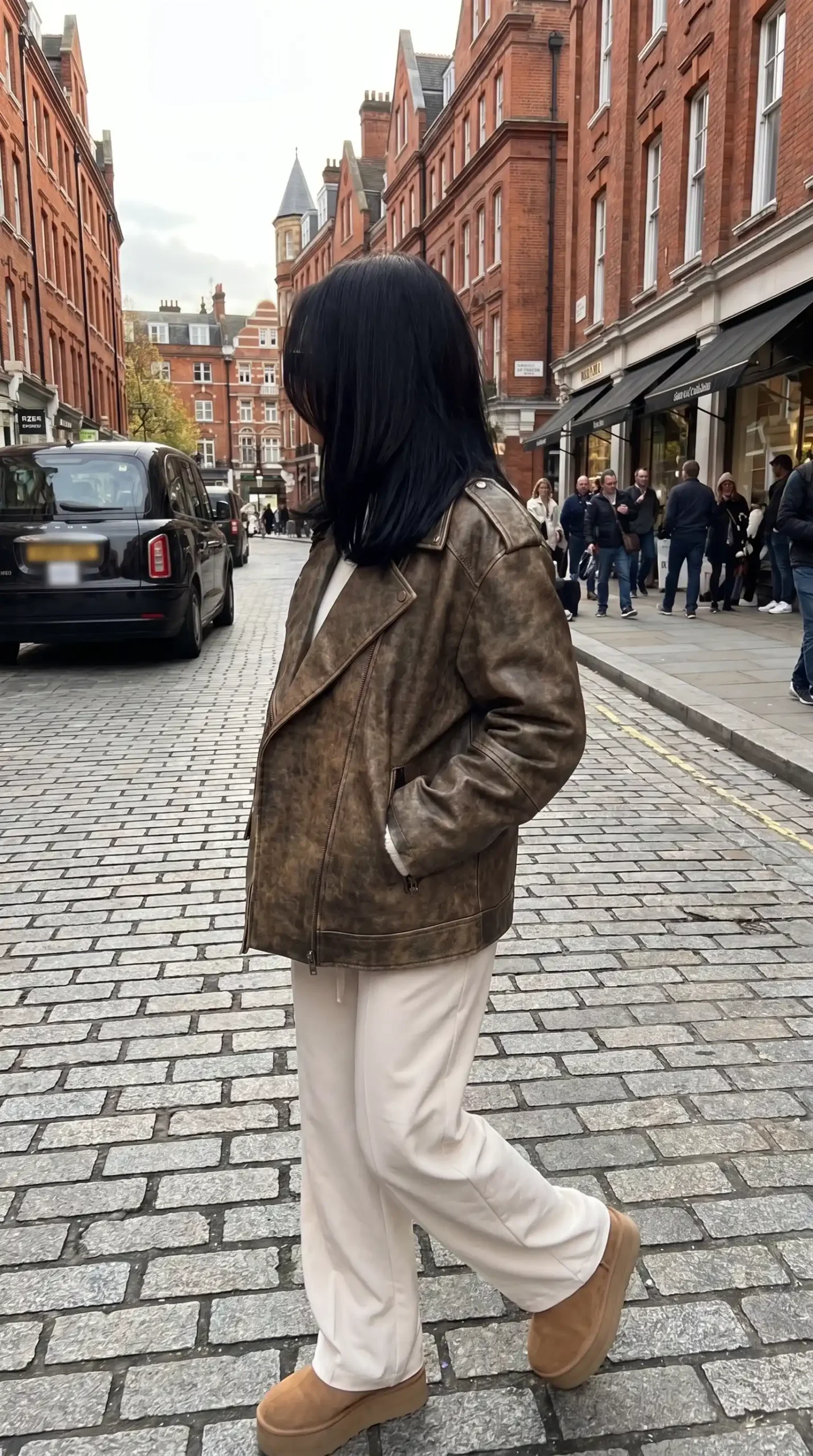 Woman wearing vintage distressed brown leather biker jacket with white wide leg trousers and tan Ugg boots on London cobblestone street