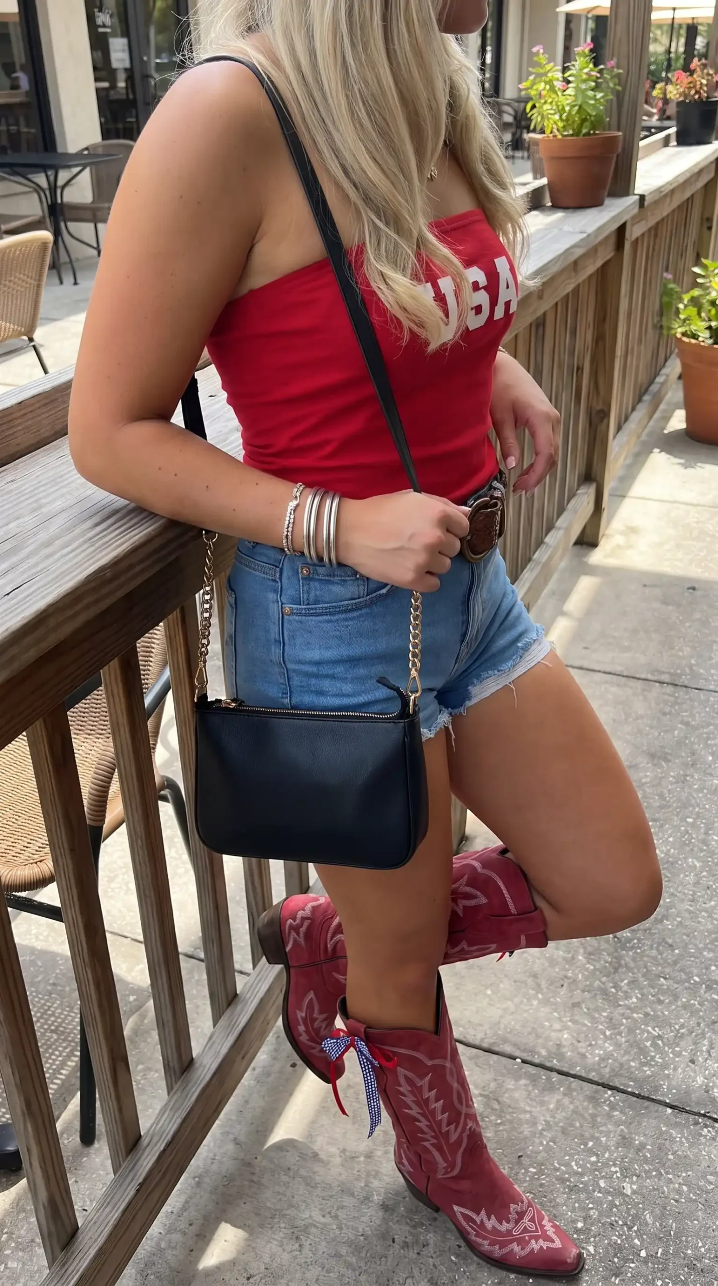 Woman wearing red USA tube top with denim cutoff shorts and red cowboy boots holding black crossbody bag
