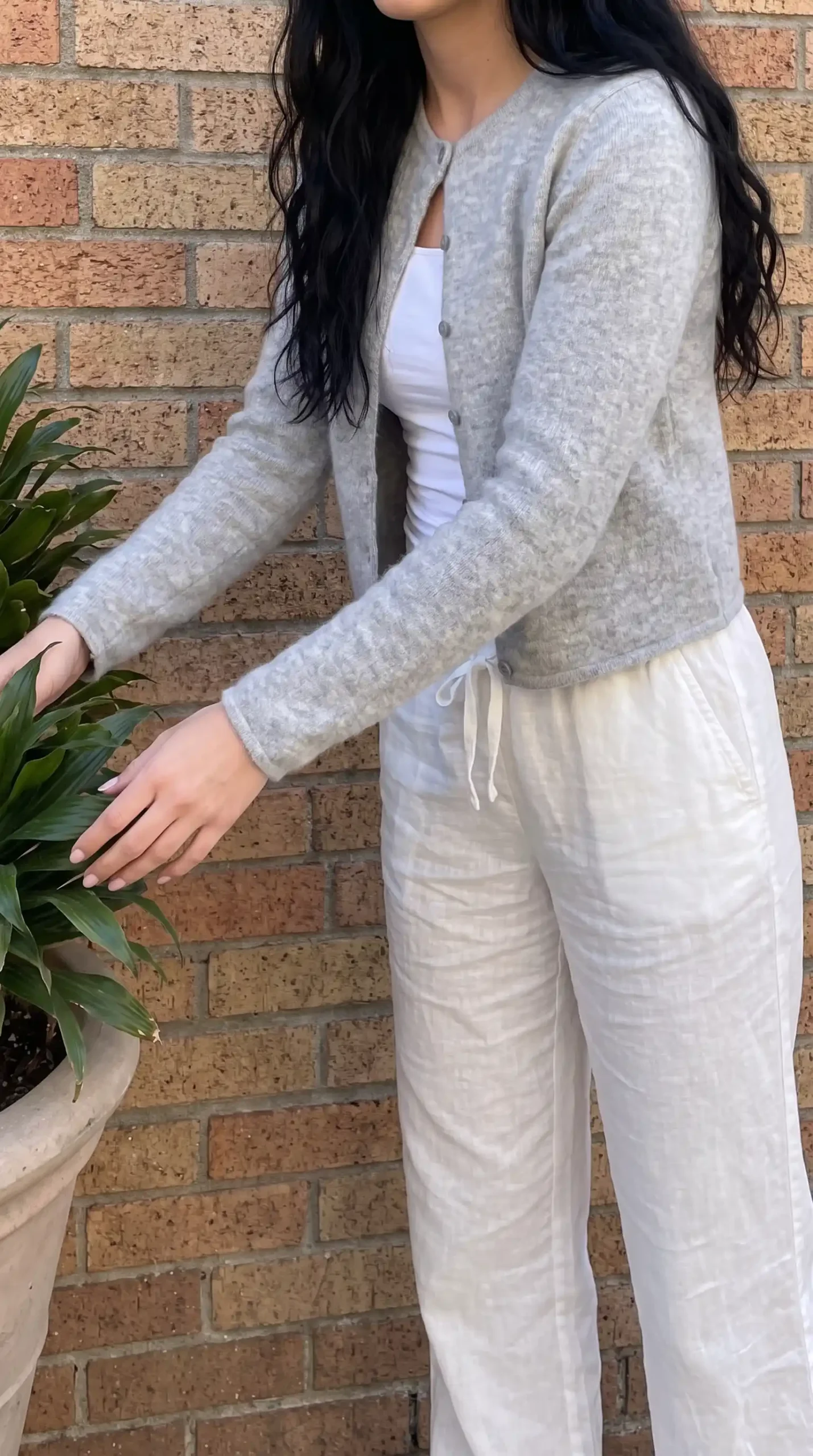 Woman wearing grey fluffy knit cardigan with white tank and cream linen trousers against brick wall