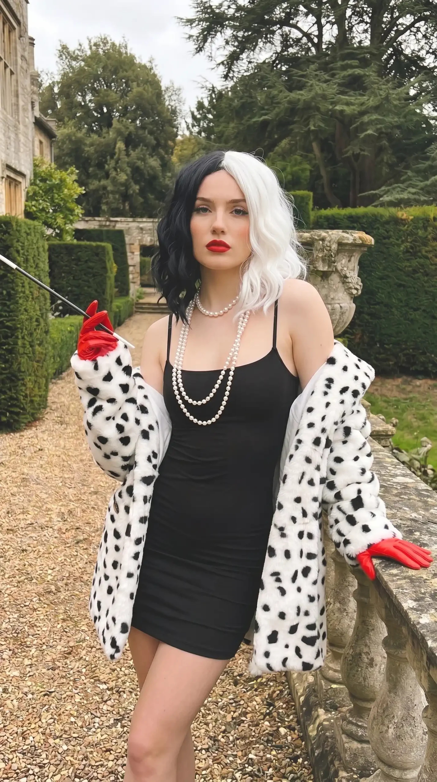 Woman dressed as Cruella de Vil in dalmatian faux fur coat black mini dress red leather gloves layered pearl necklaces and half black half white wig