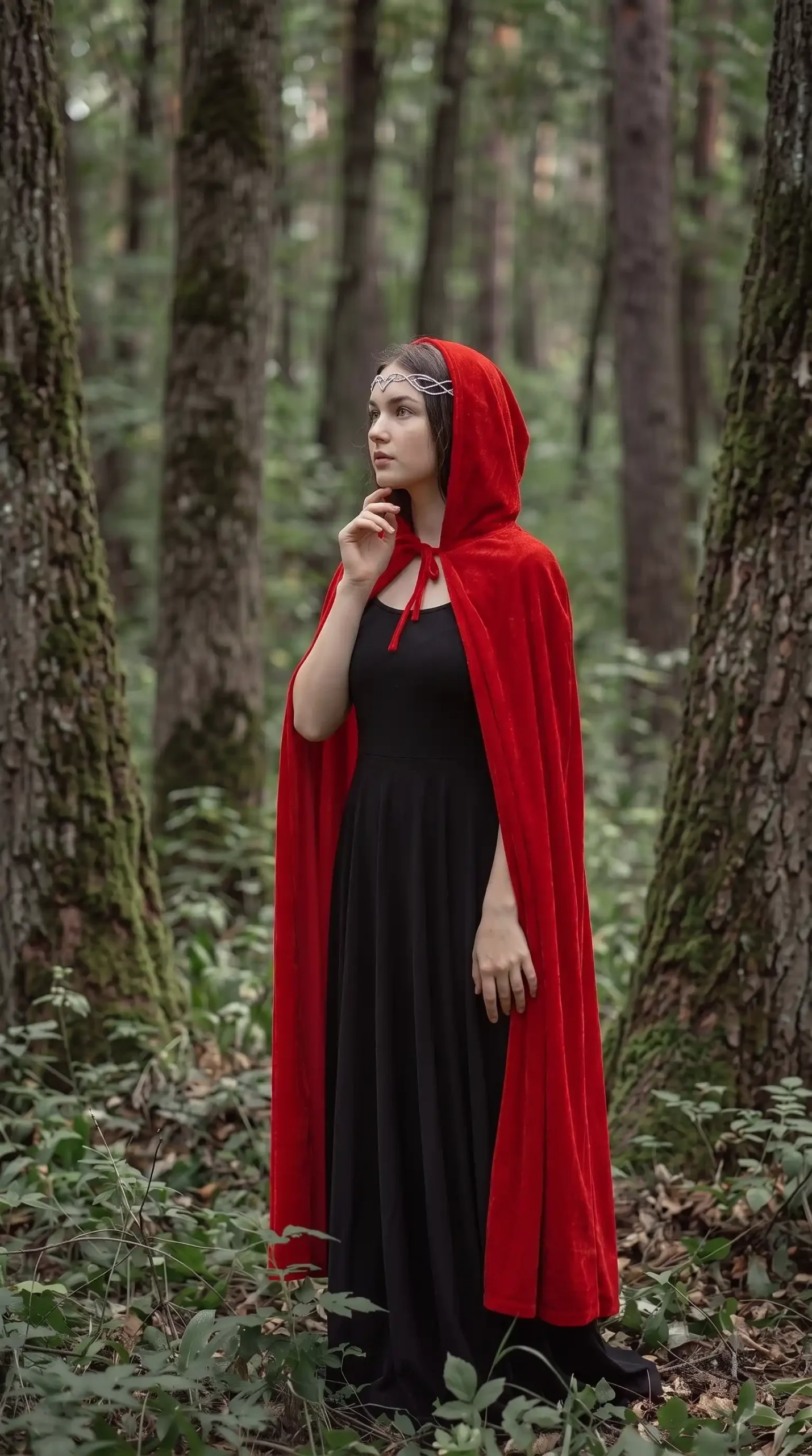 Woman dressed as Little Red Riding Hood in red velvet hooded cape over black maxi dress with silver headpiece in forest