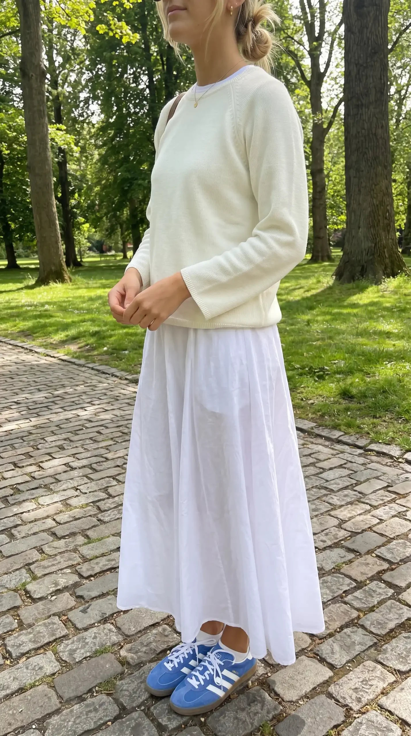 Woman wearing cream knit sweater with white flowy maxi skirt and blue Adidas Gazelle sneakers in park