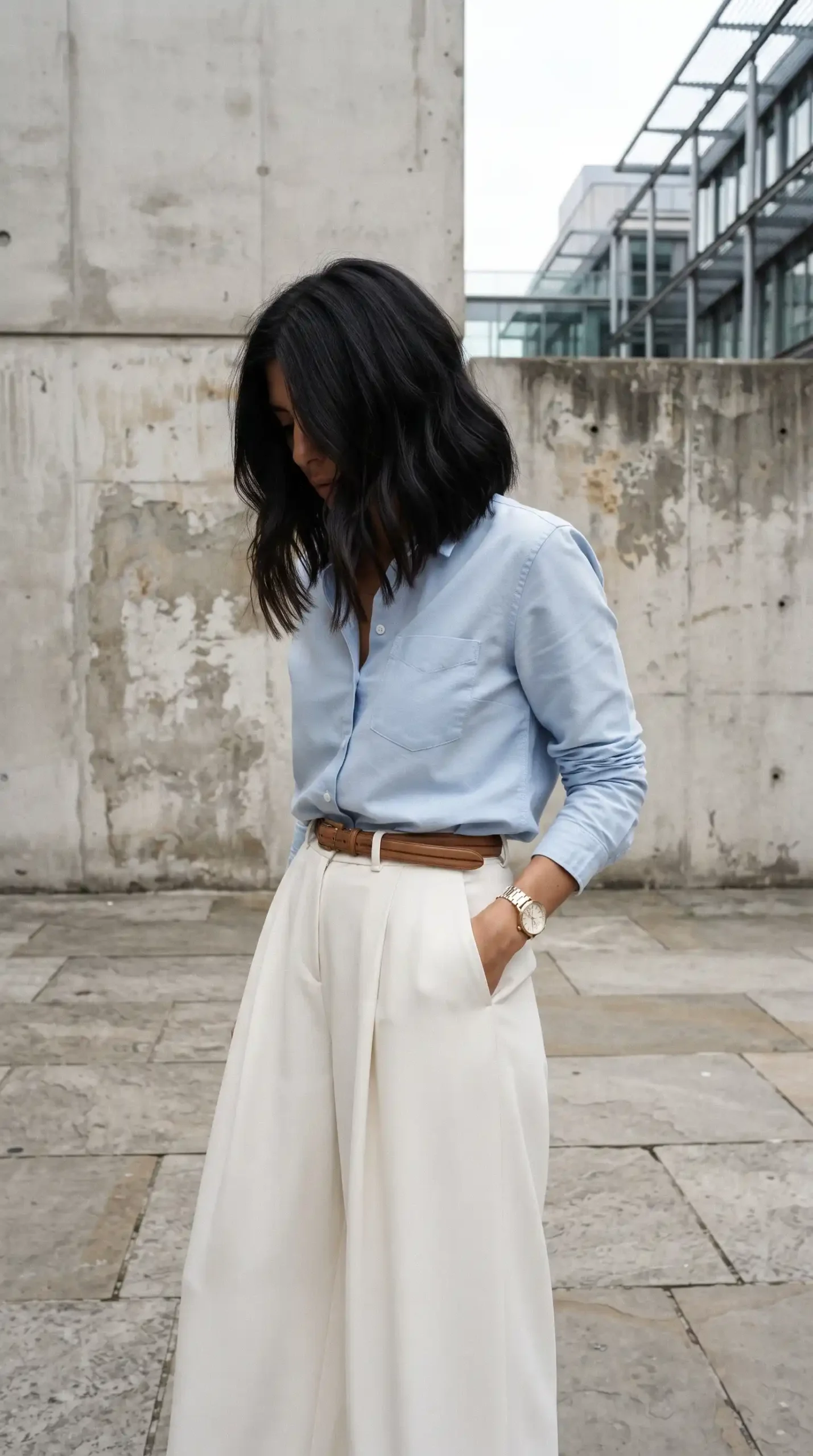 Woman wearing light blue oxford shirt with cream wide leg pleated trousers and tan leather belt outdoors