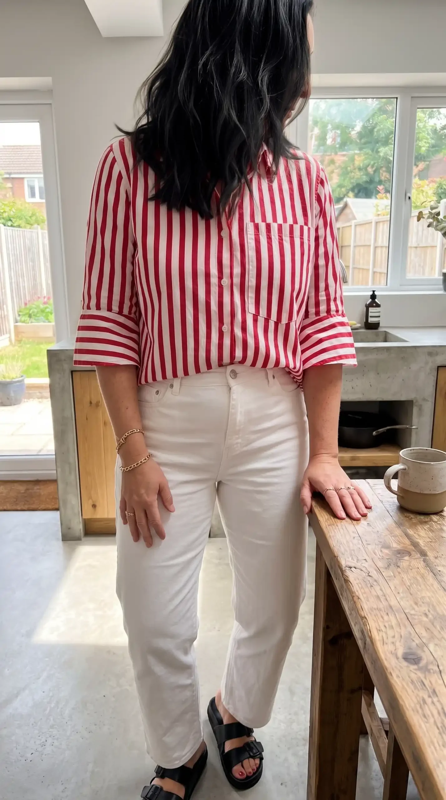 Woman wearing red and white striped oversized cropped shirt with white straight leg jeans and black chunky sandals indoors