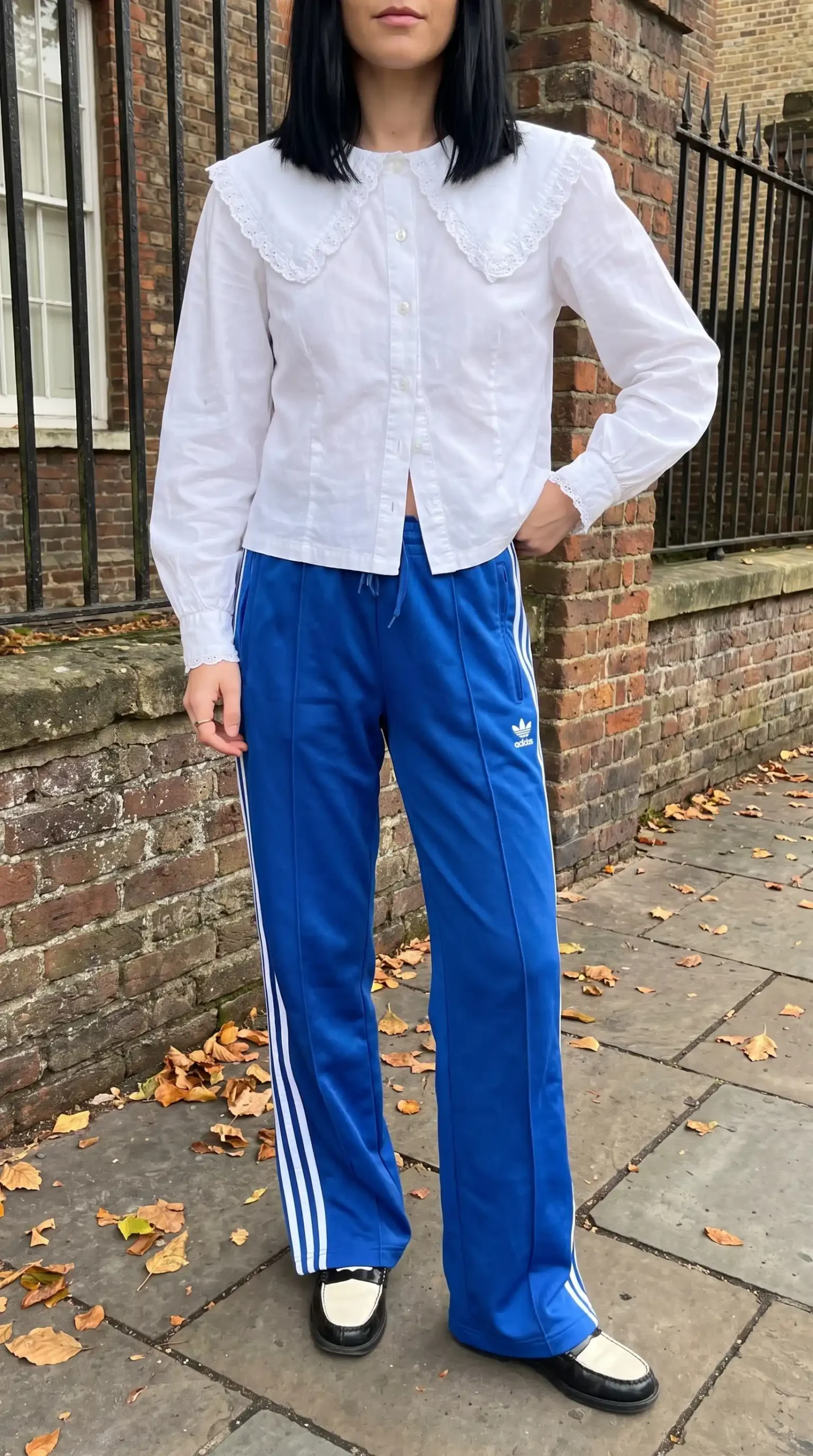 Woman wearing white lace Peter Pan collar blouse with blue Adidas track pants and black white loafers on London street