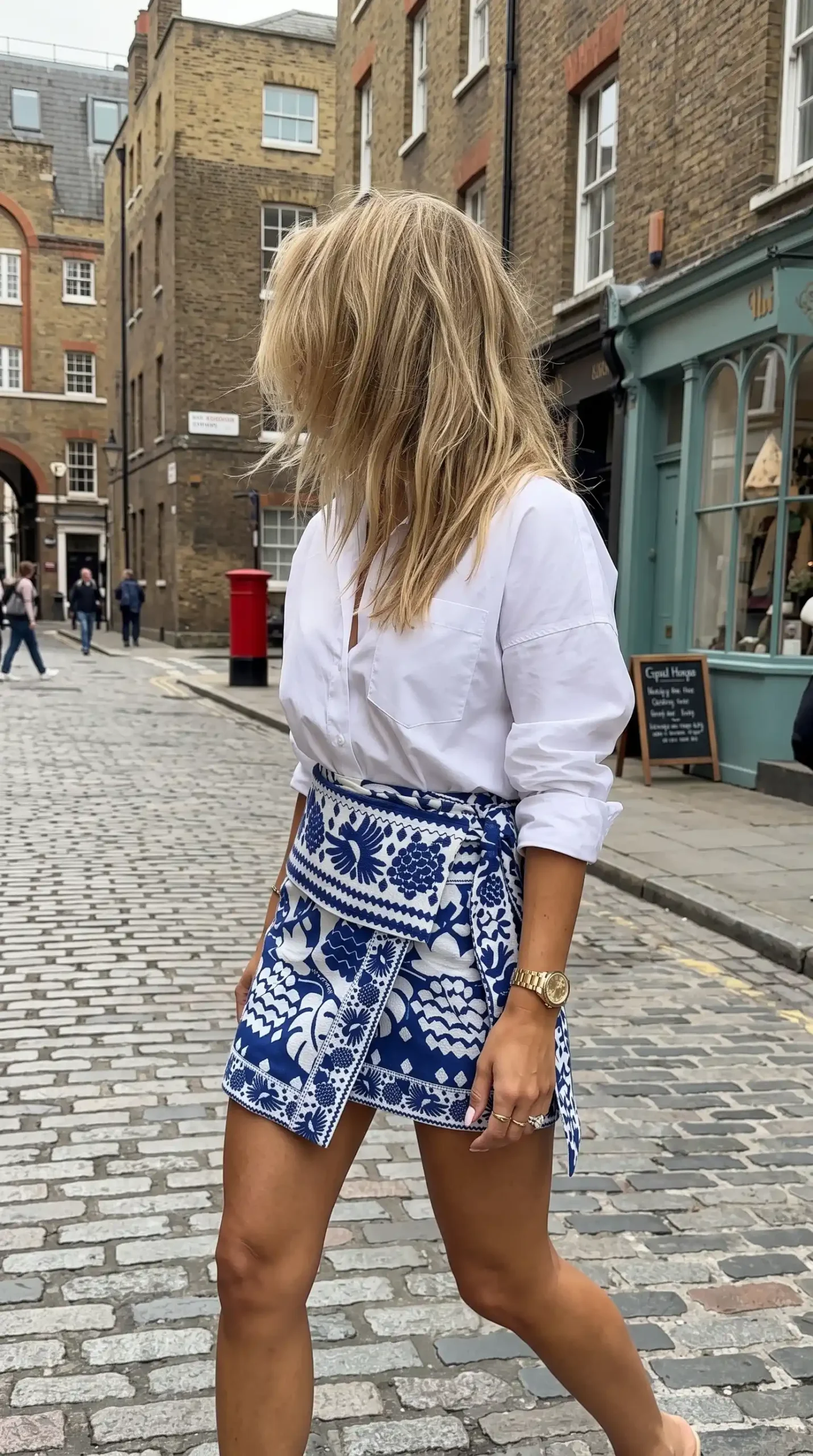 Woman wearing white oversized button down shirt with blue and white floral print wrap mini skirt on London cobblestone street