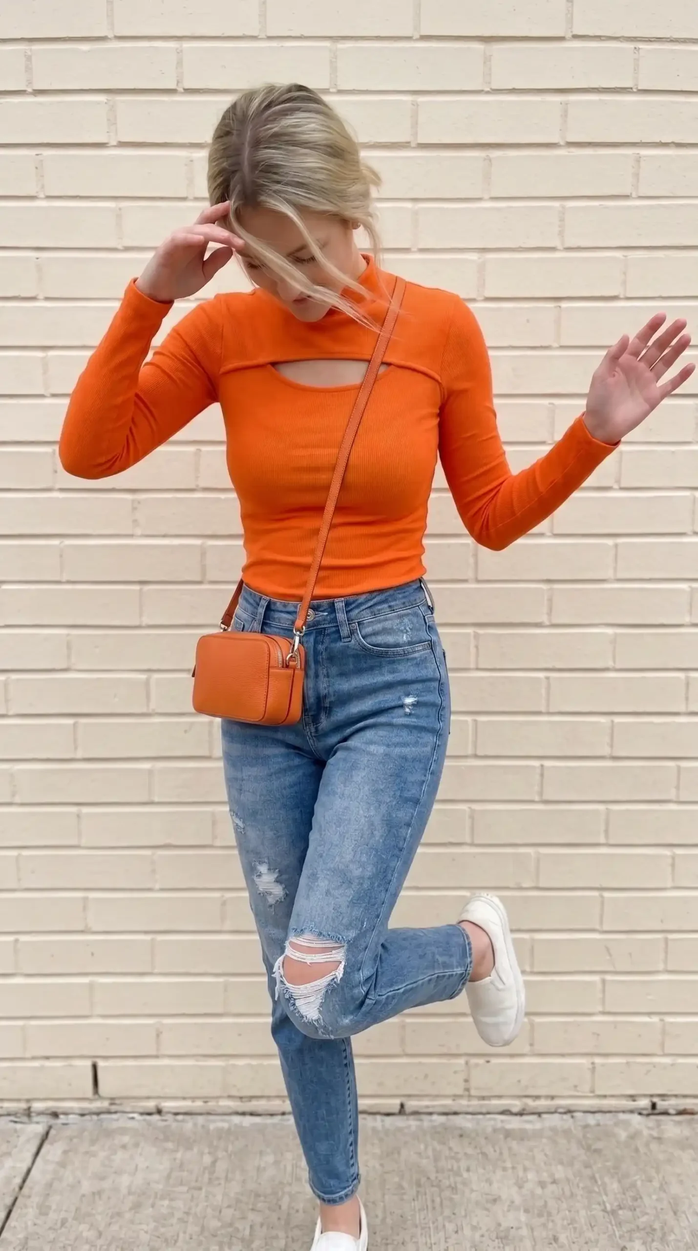 Woman wearing orange ribbed mock neck cutout long sleeve top with ripped mid wash mom jeans and white sneakers