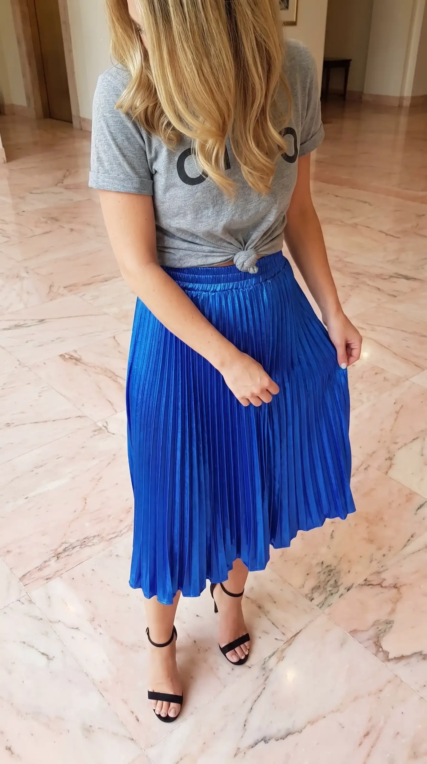 Woman in grey knotted graphic tee with royal blue metallic pleated midi skirt and black heels