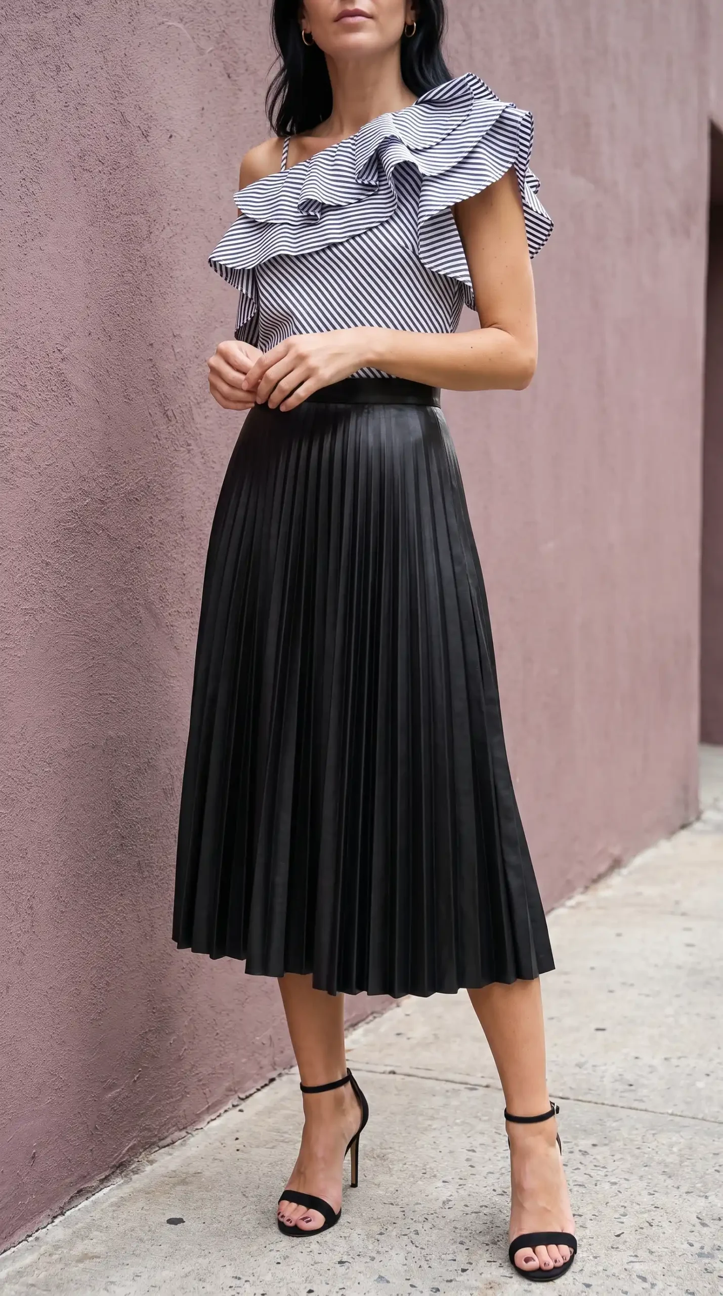 Woman in navy striped one shoulder ruffle top with black faux leather pleated midi skirt and black heels