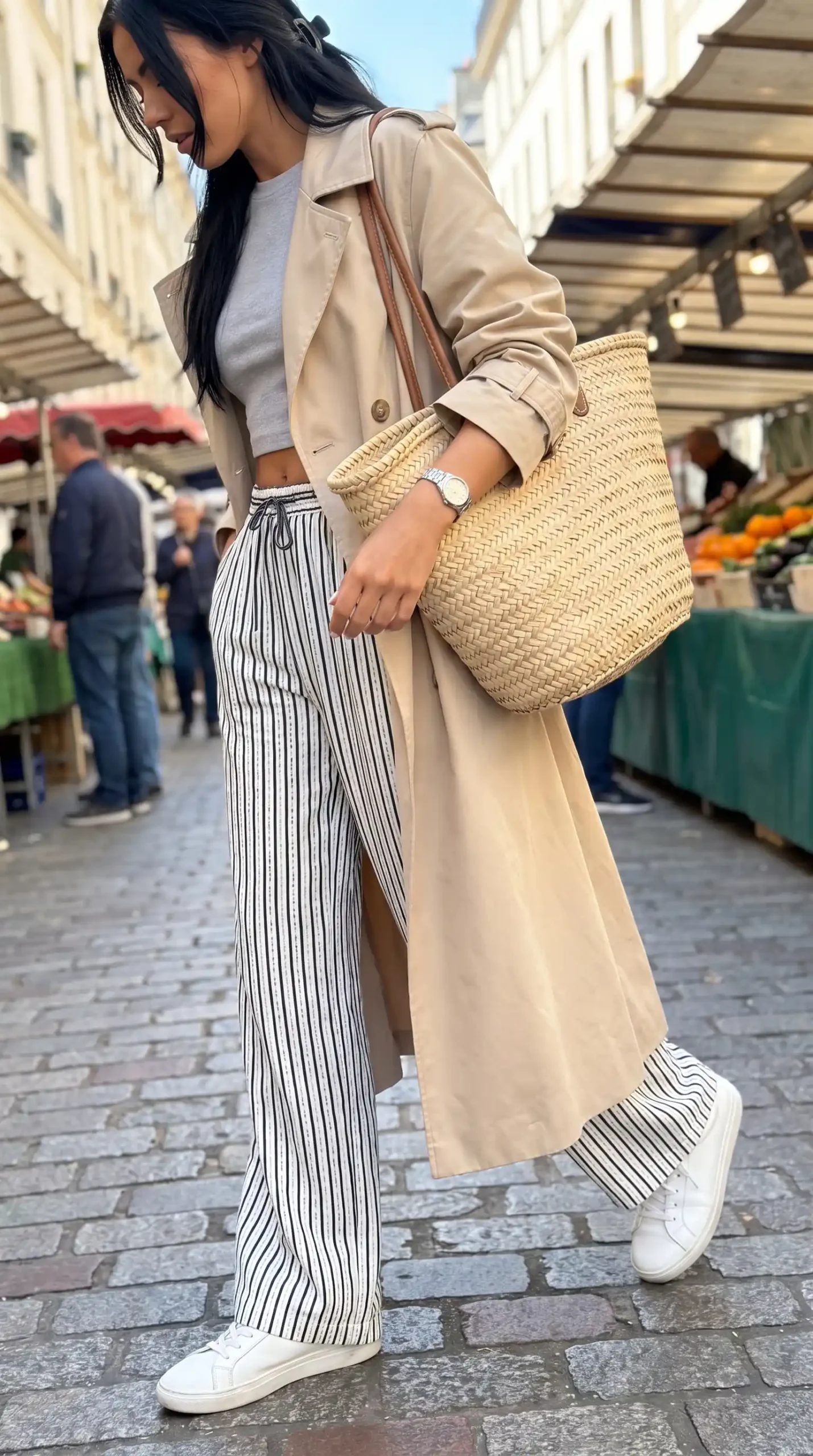 Woman wearing beige trench coat with grey crop top black and white striped wide leg trousers and white sneakers outdoors