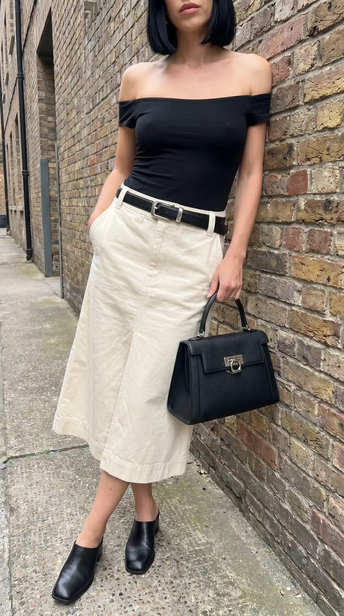 Woman wearing black off shoulder top with cream midi skirt black belt and black mules outdoors