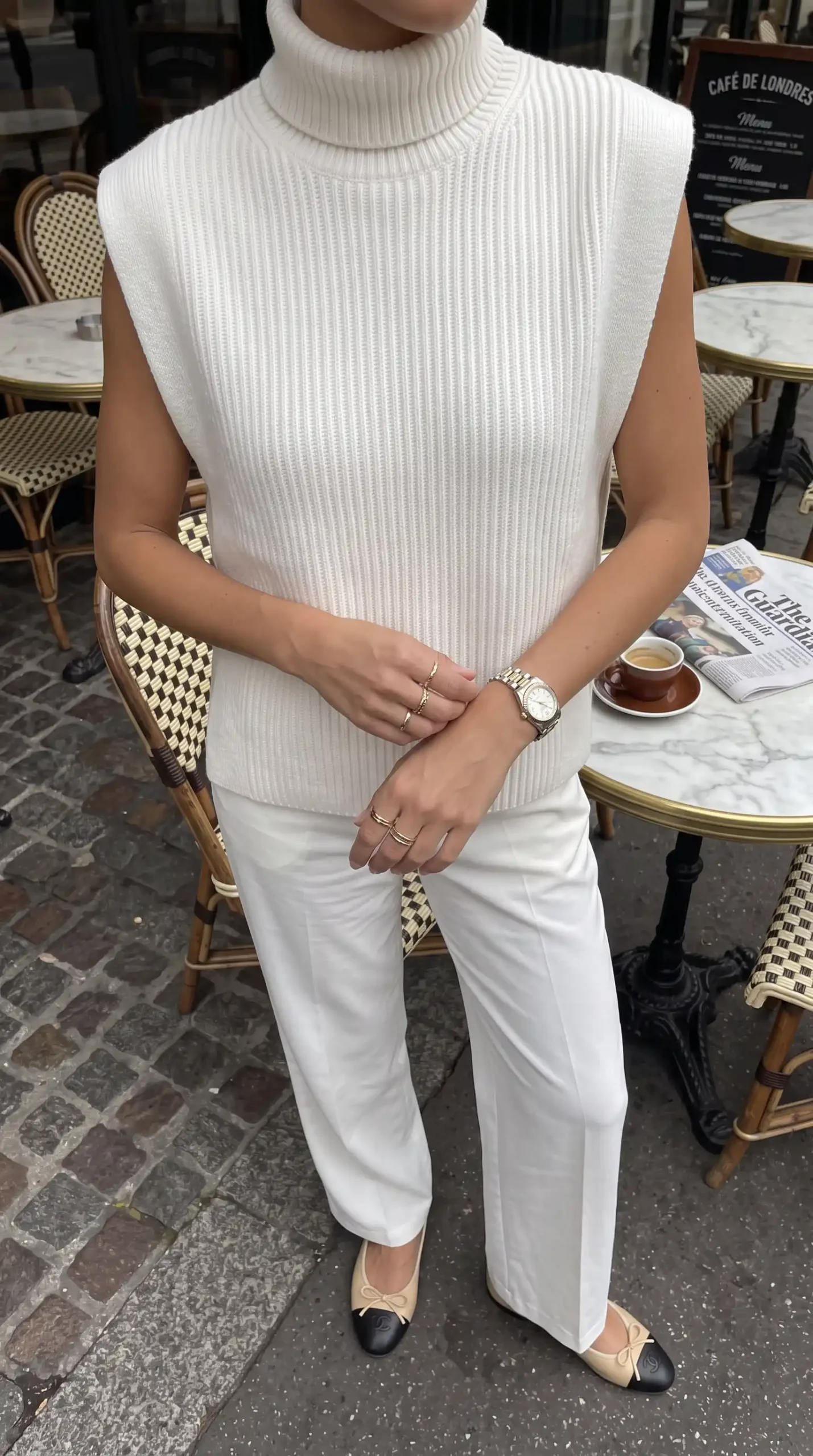 Woman wearing cream ribbed sleeveless turtleneck knit vest with white wide leg trousers and two tone ballet flats outdoors