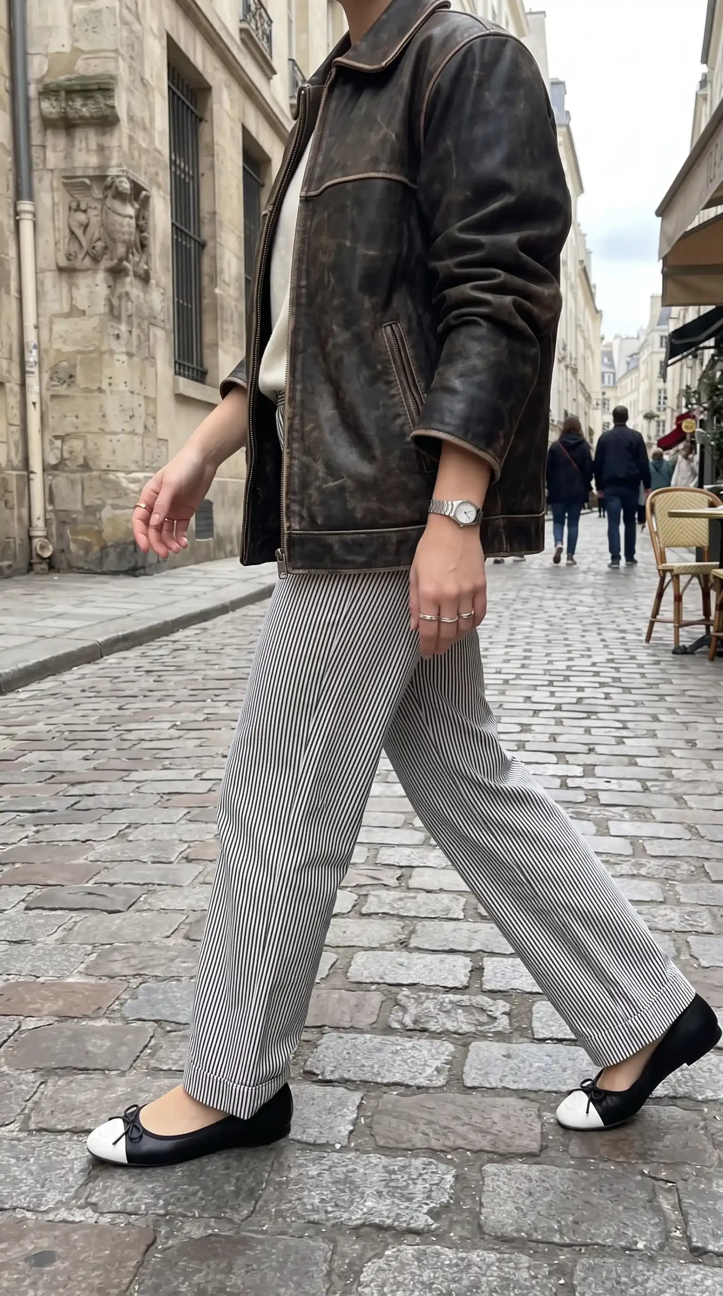 Woman wearing distressed dark brown leather jacket with black and white striped wide leg trousers and two tone ballet flats outdoors