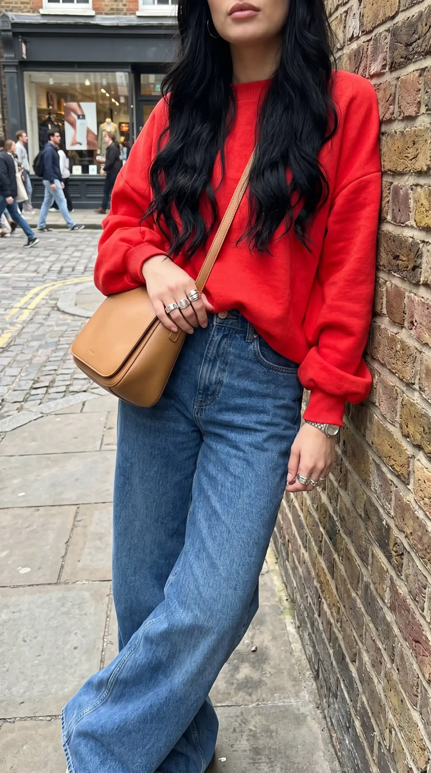 Woman wearing red oversized crewneck sweatshirt with mid wash wide leg jeans and tan crossbody bag outdoors