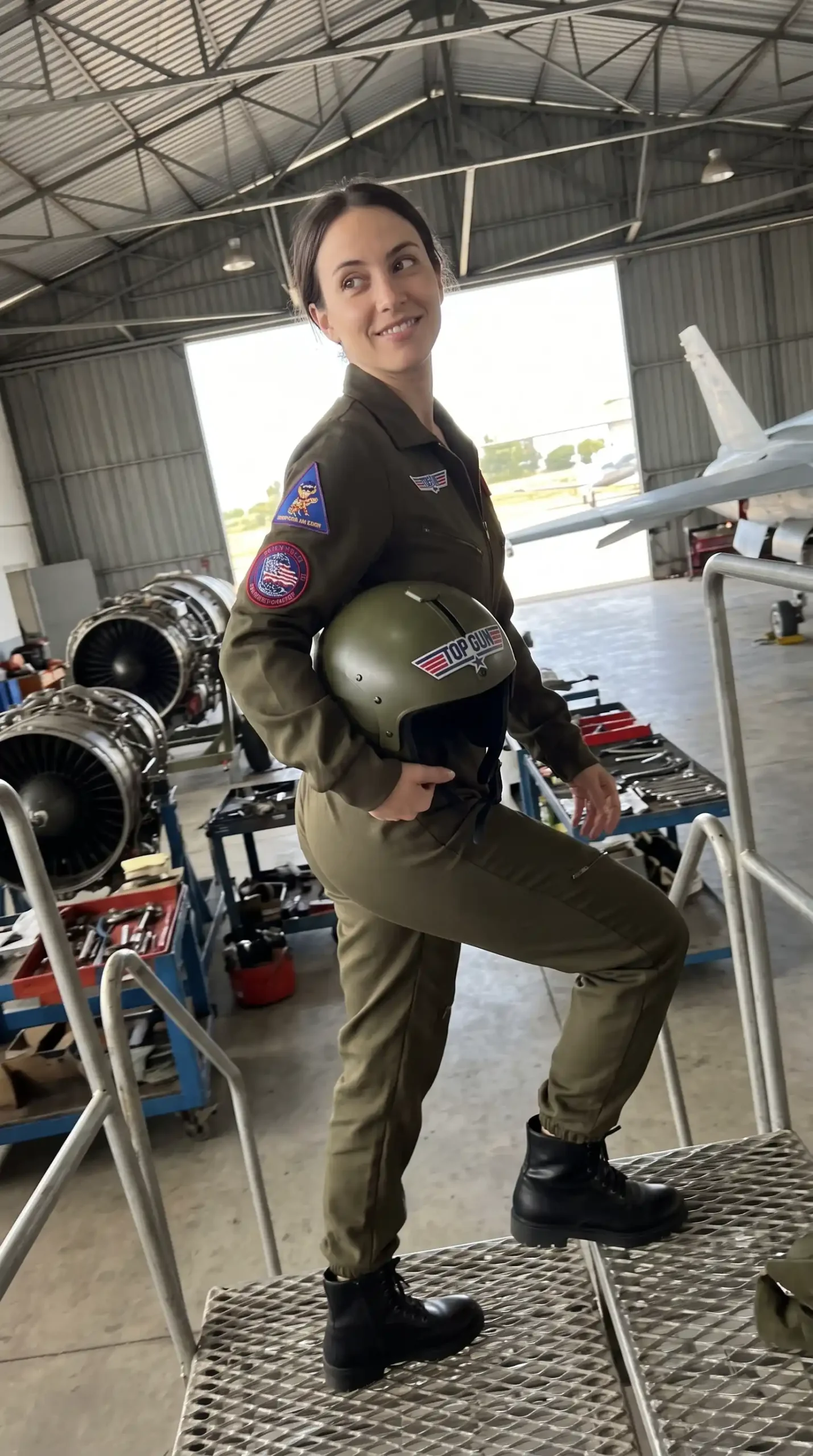 Woman in Top Gun olive green flight suit with patches and pilot helmet in aircraft hangar