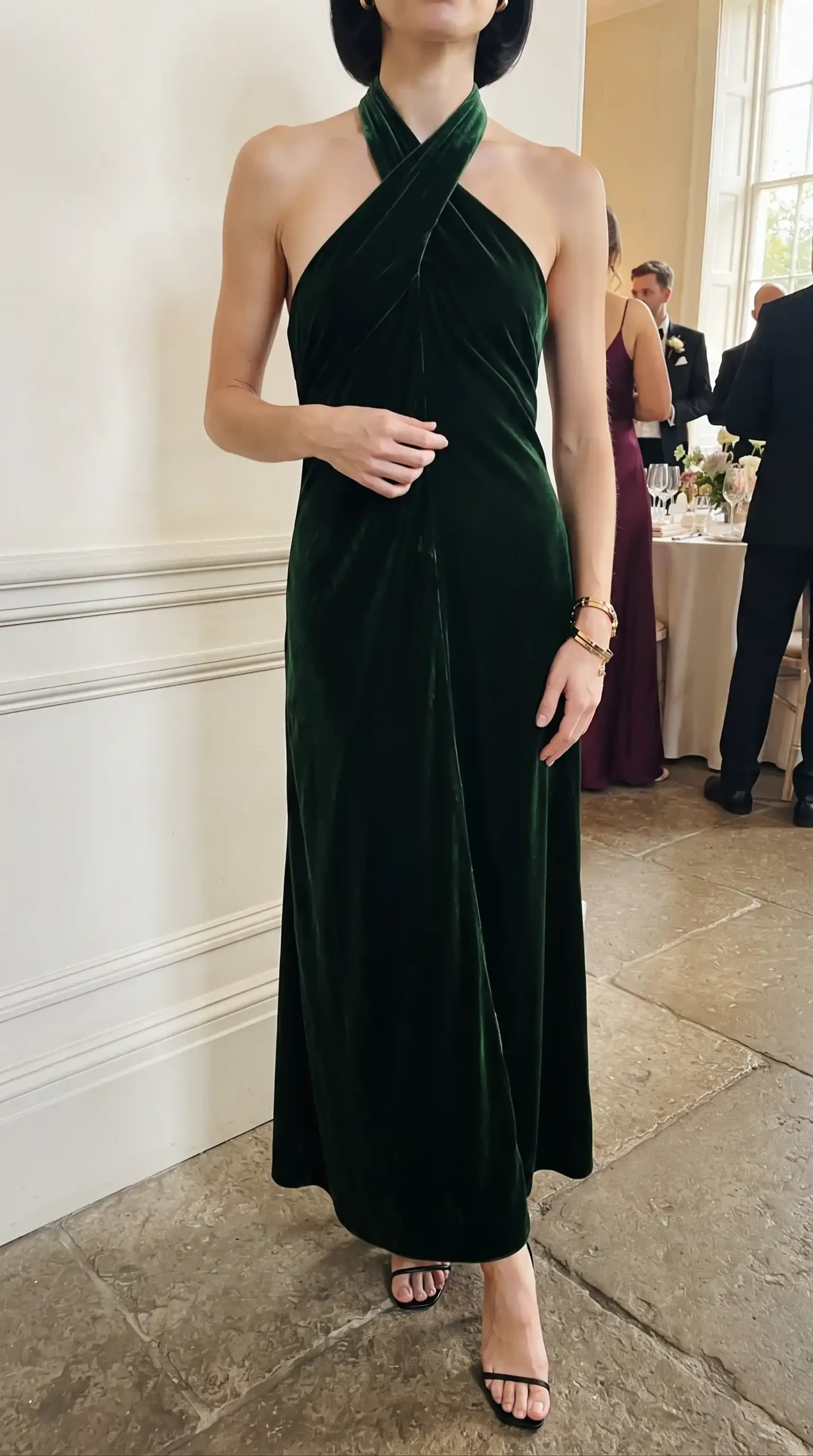Woman wearing emerald green velvet cross halter neck sleeveless maxi dress with black heels at wedding venue