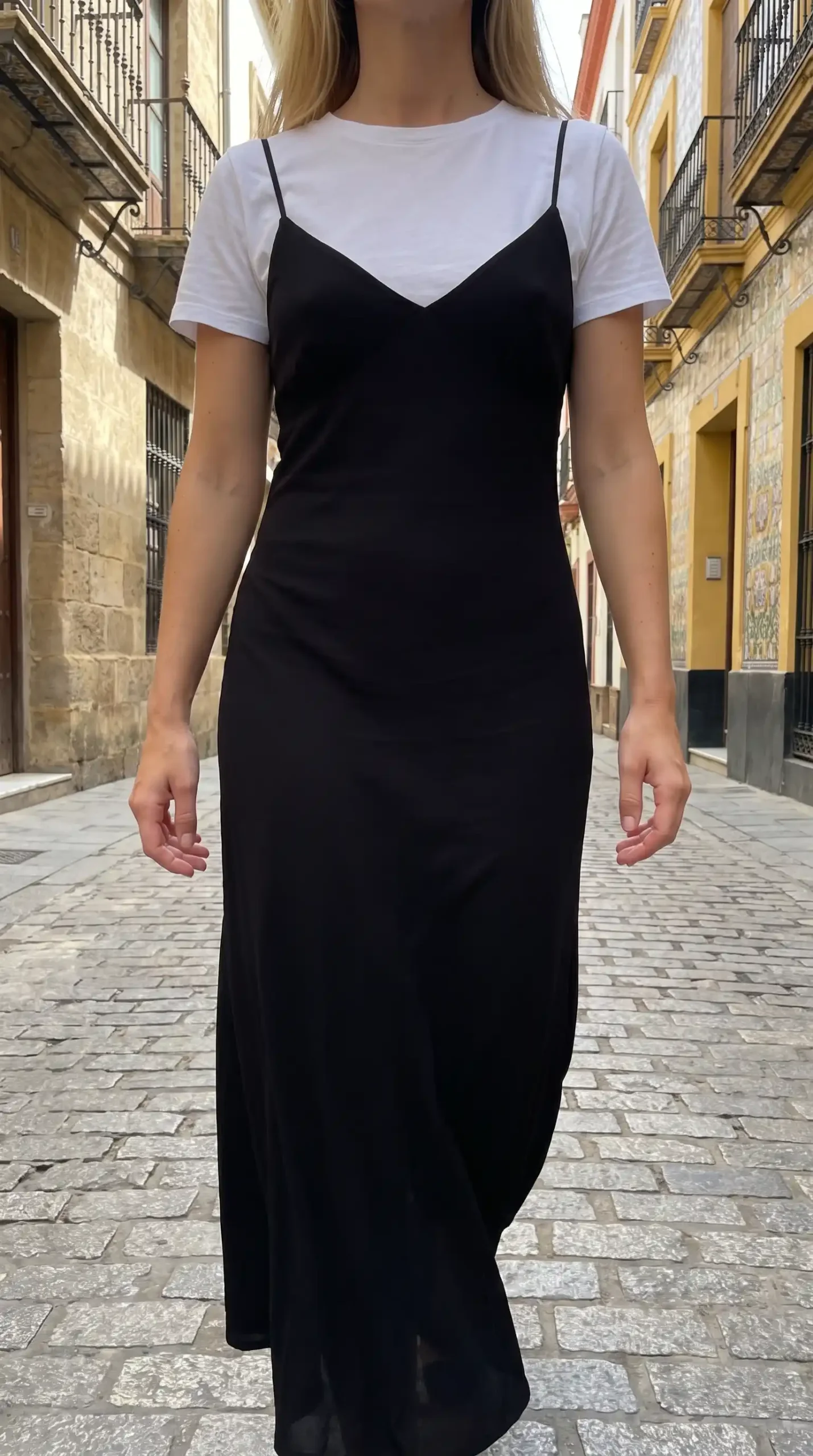 Woman wearing white t-shirt layered under black satin slip maxi dress on cobblestone street