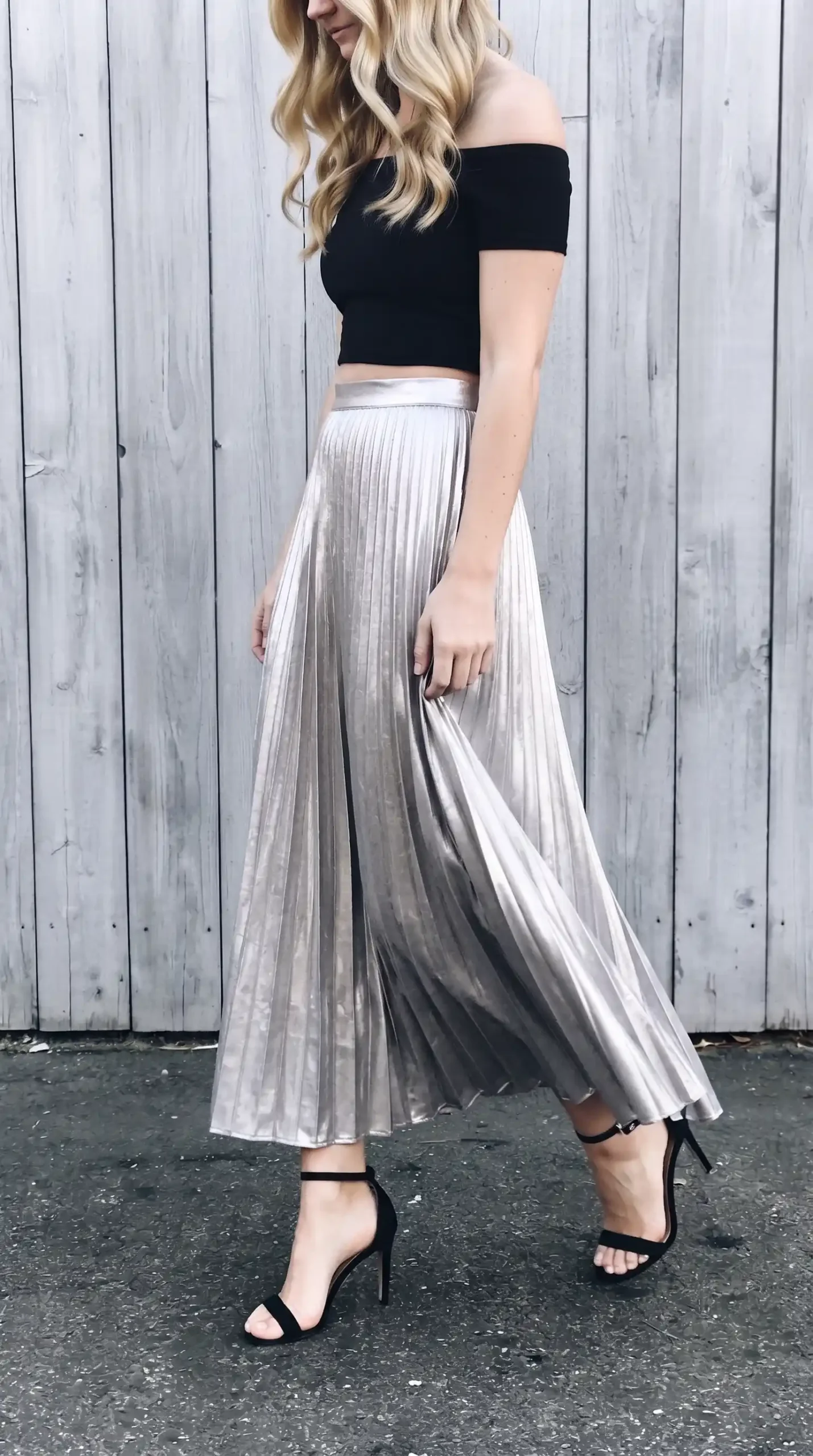 Woman in black off shoulder crop top with silver metallic pleated maxi skirt and black strappy heels